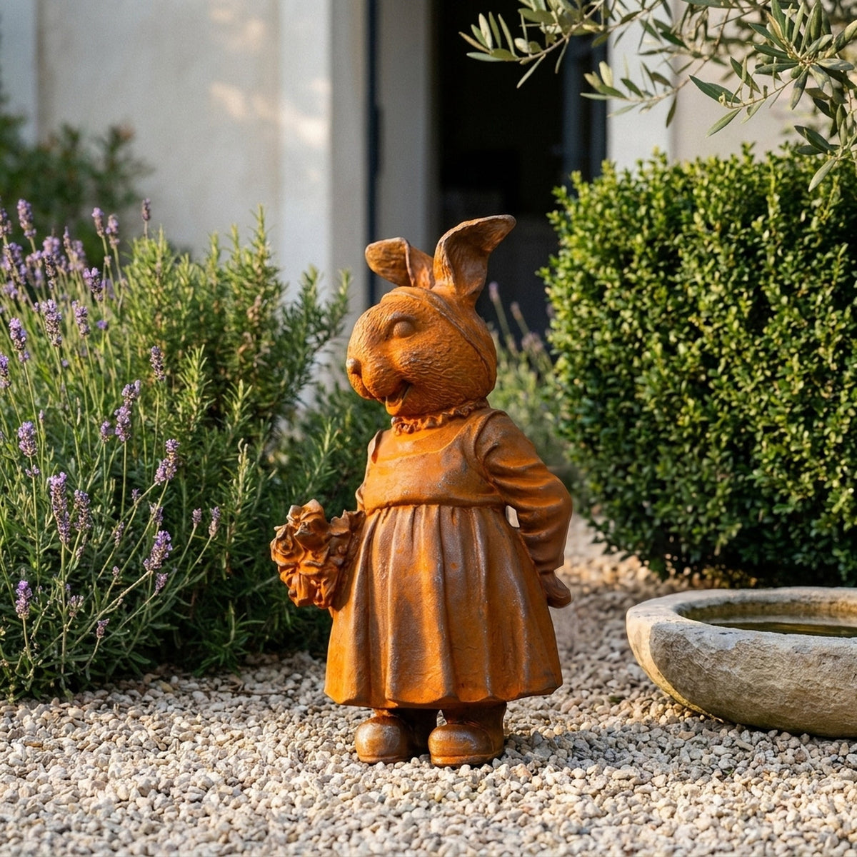 Miss Clover rust cast iron rabbit statue in a pinafore holding a posy, on gravel beside lavender and a stone water basin