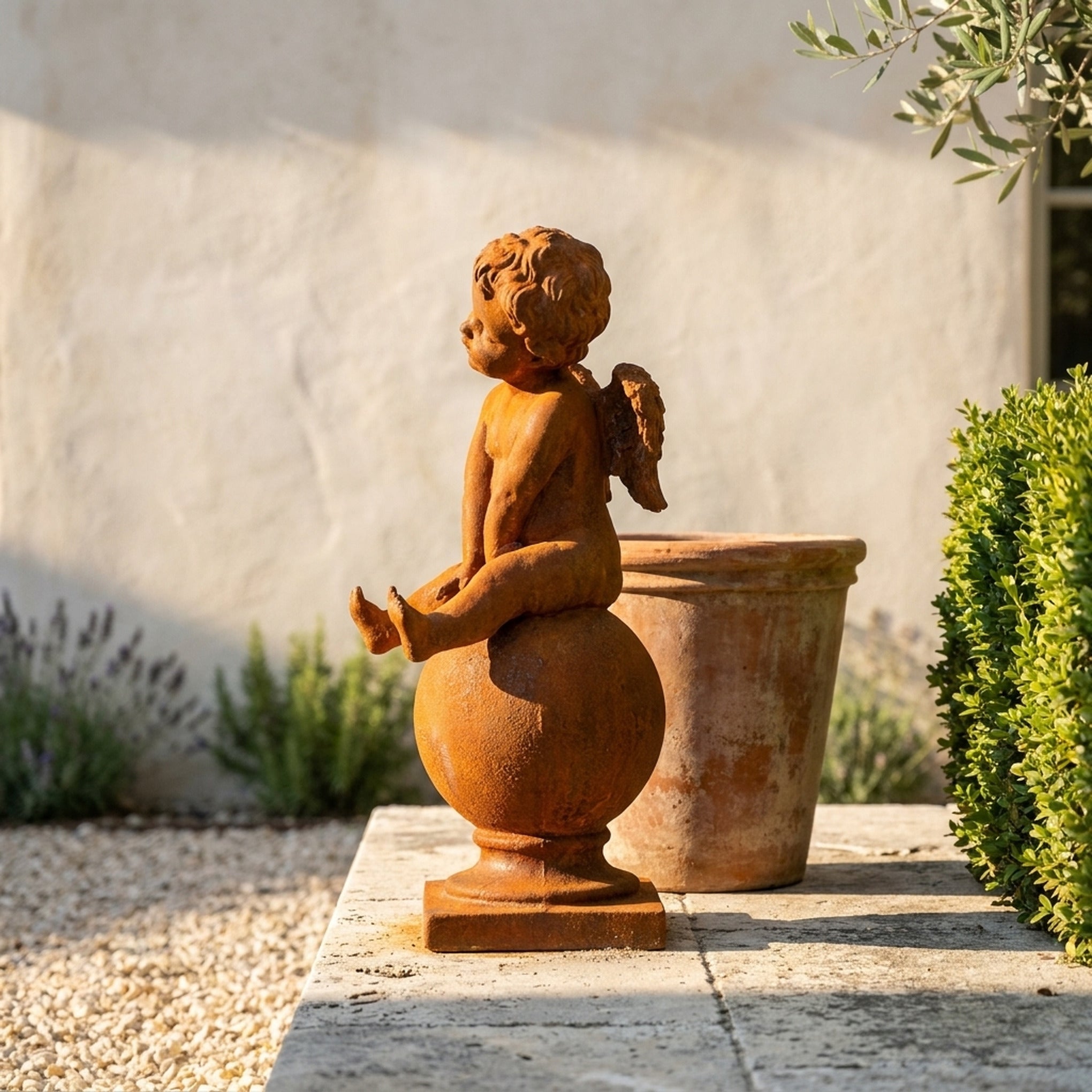 Hugo Wingsley rust cast iron cherub statue seated on a sphere, standing on gravel beside a terracotta pot