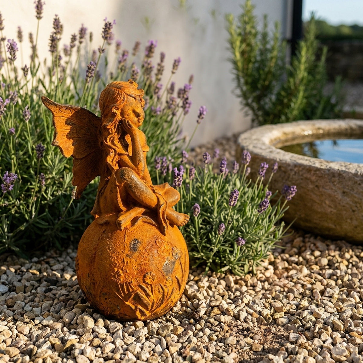 Lady Ophelia rust cast iron fairy statue seated on a flowered sphere, on gravel beside lavender and a stone birdbath