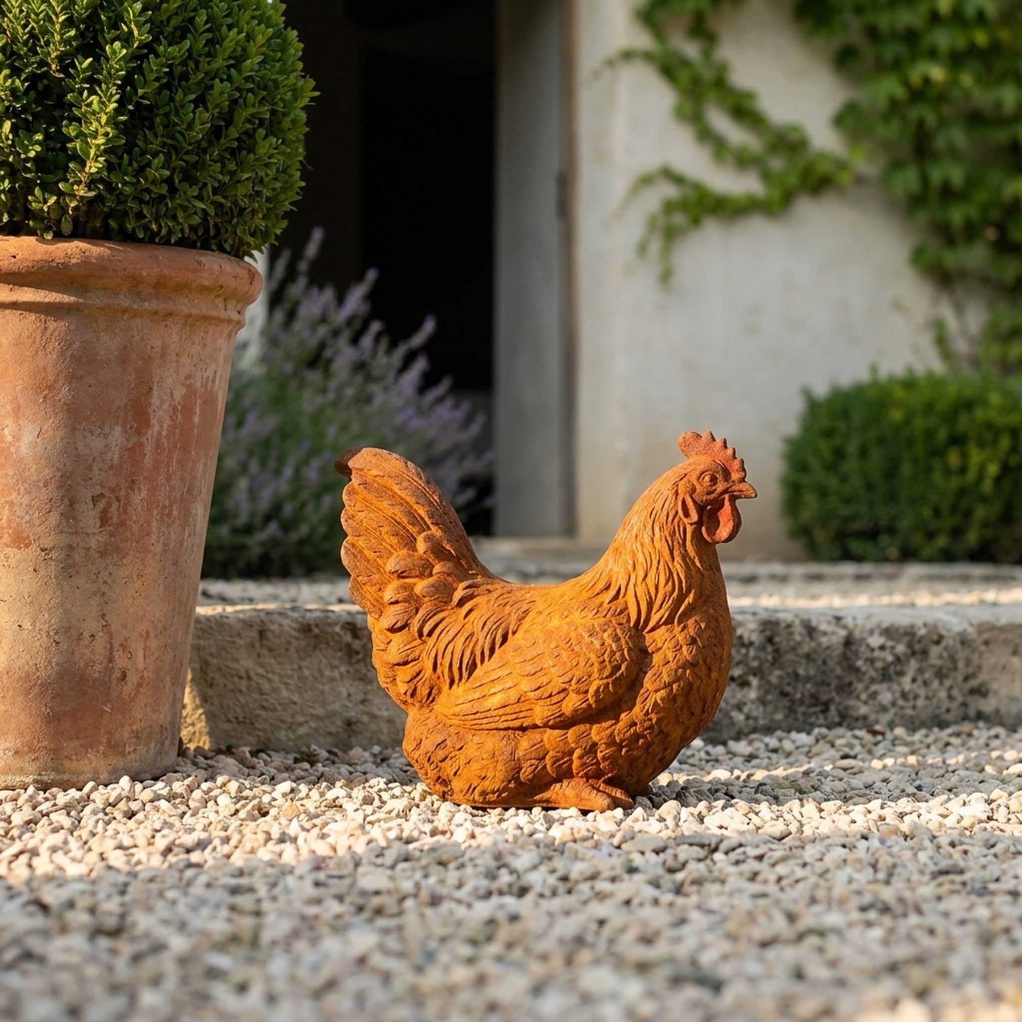 Dame Henrietta rust cast iron nesting hen statue, sitting on gravel beside a terracotta pot