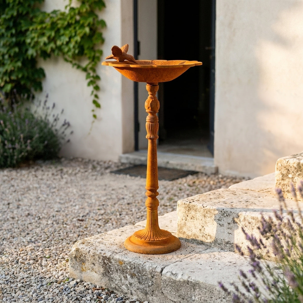 The Willowmere rust cast iron pedestal bird feeder with a bird on the rim, standing on gravel in a courtyard