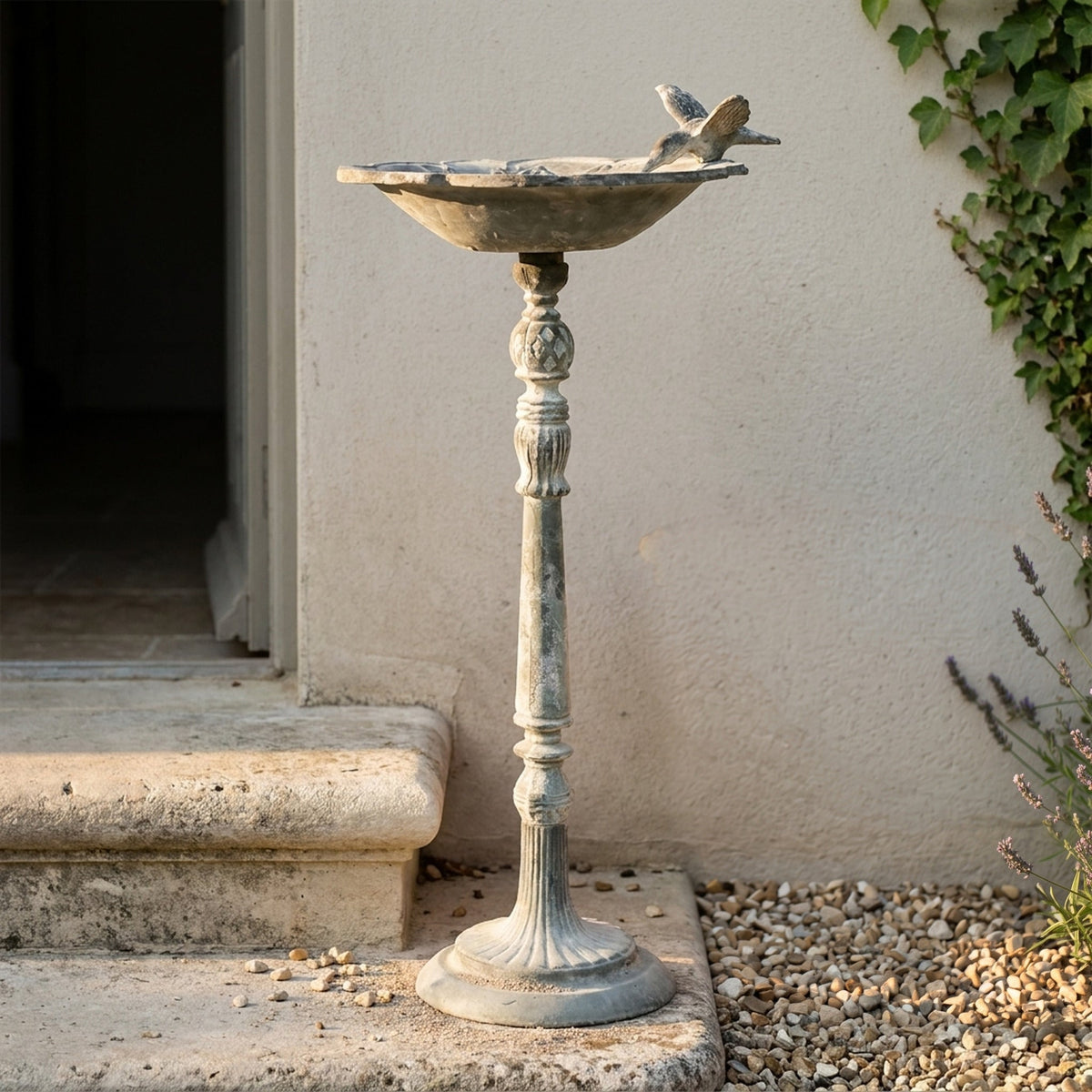 The Willowmere in weathered grey cast iron, a pedestal bird feeder with a bird on the rim, on gravel in a courtyard