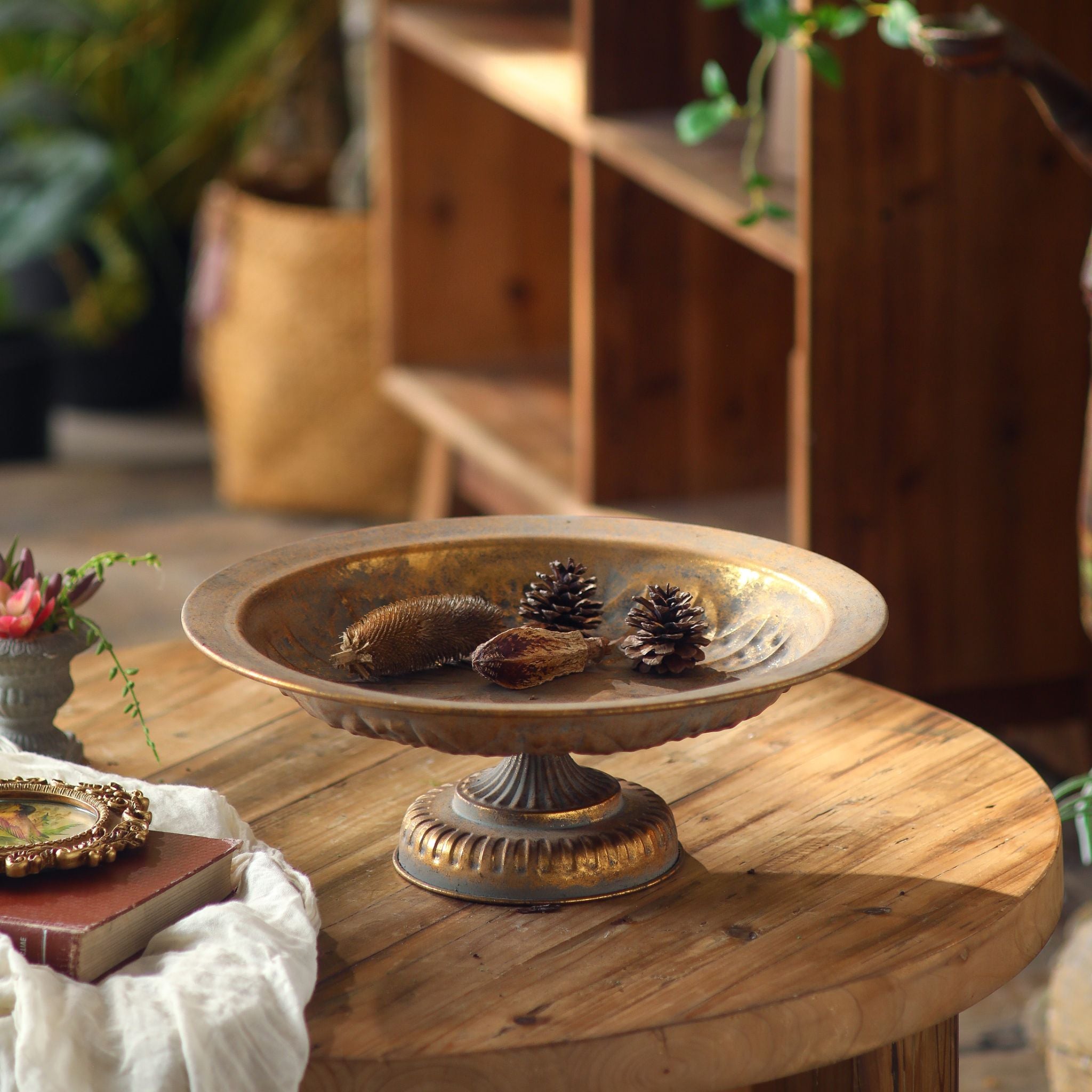 Menton pedestal bowl in burnished gold styled with pine cones on a round timber table. Hero.