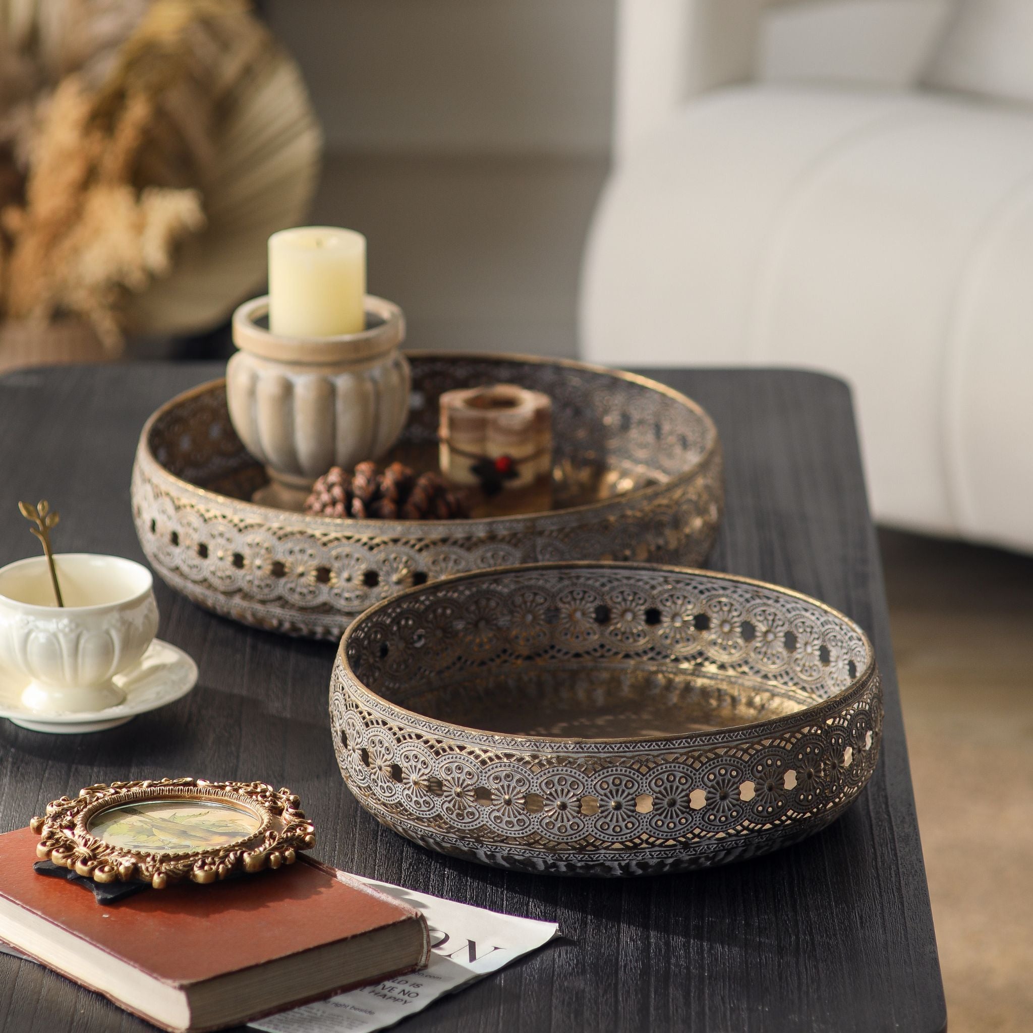 Abrielle set of two French Provincial metal trays with pierced lace edge and antique gold whitewashed finish, styled on a dark coffee table with a candle and pinecones