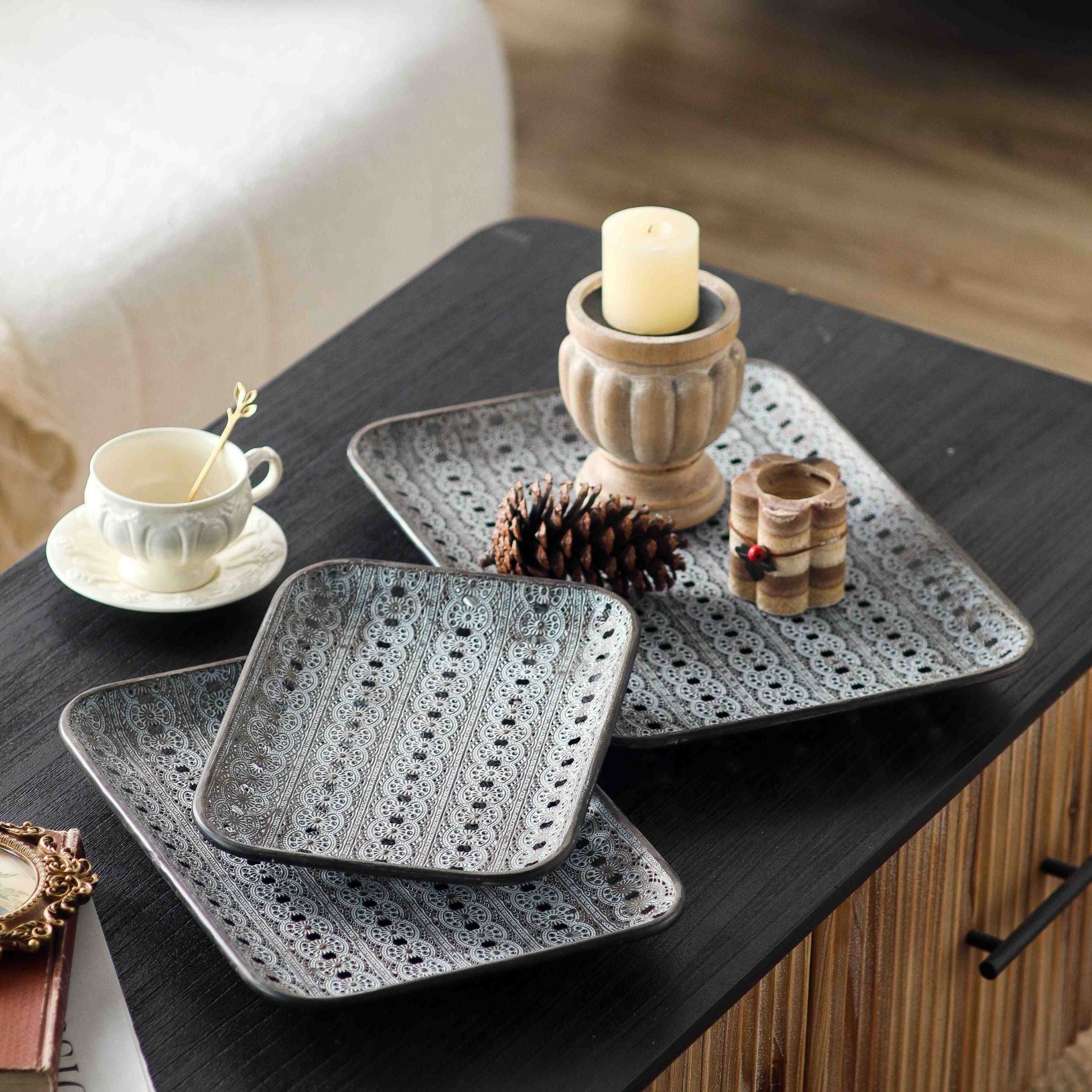 Adélaïde set of three French Provincial metal trays with a pierced lace pattern and whitewashed grey finish, styled on a dark coffee table with a candle and pinecone