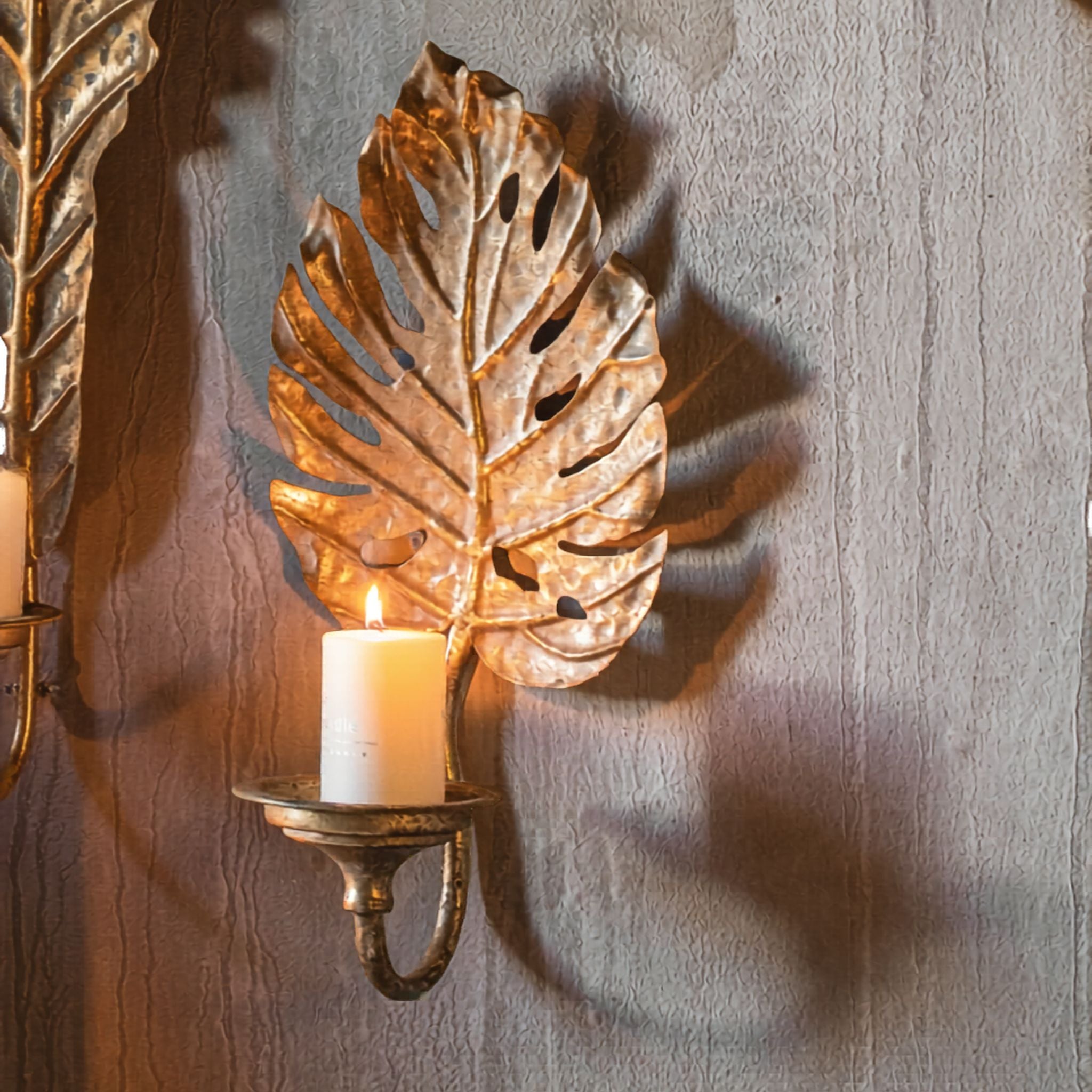 Adele French Provincial split-leaf monstera wall sconce in antique gold aged metal, with a scrolled arm holding a lit pillar candle, mounted on a wall. Hero.