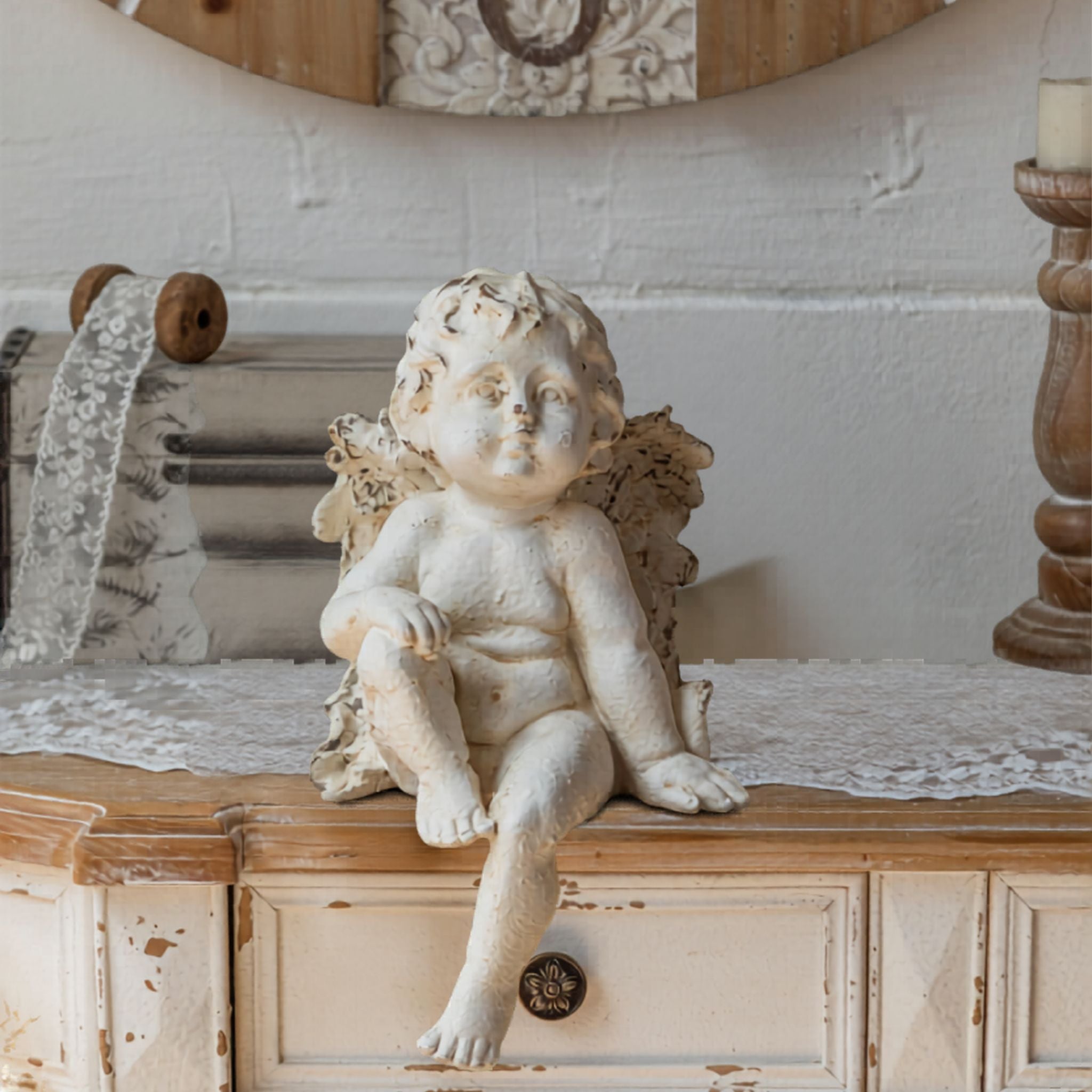 Antoine French Provincial cherub figurine in distressed whitewash, seated on a wooden console with one leg dangling, head tilted upward. Hero.