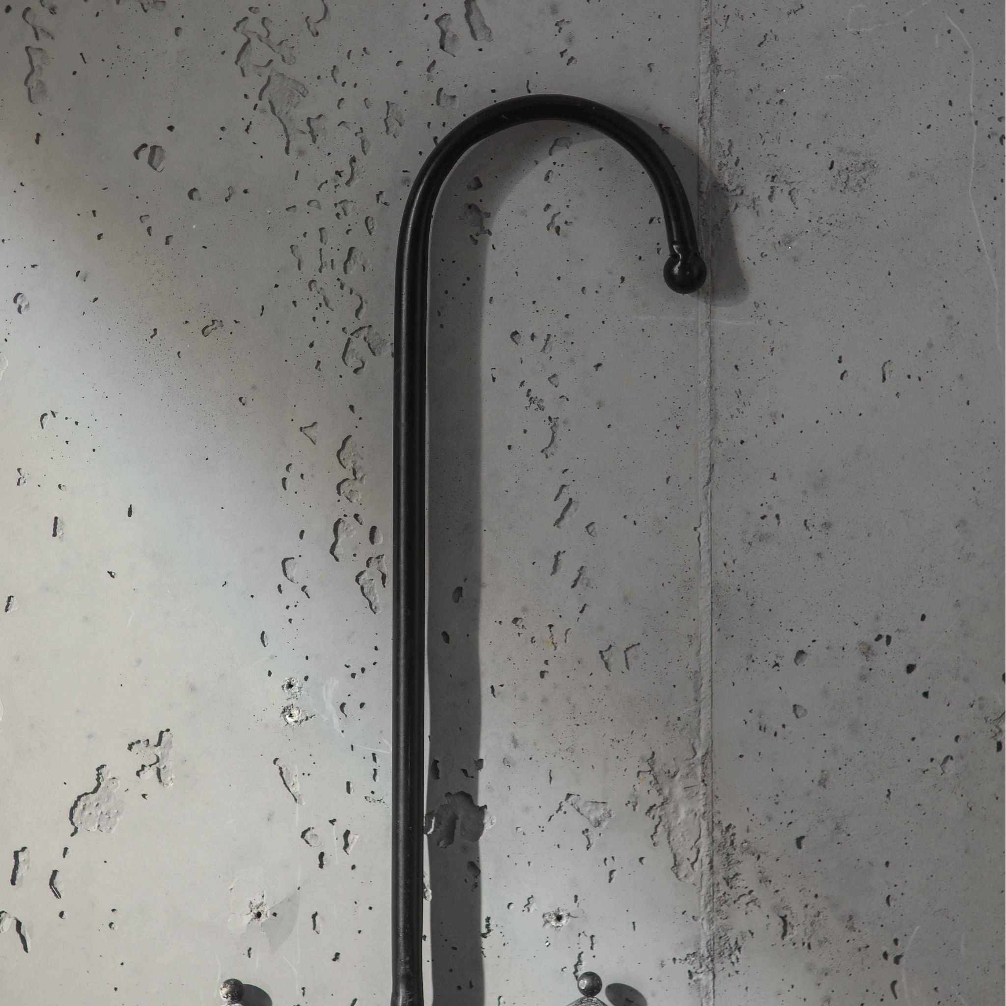 Close-up of the black curved iron hook handle of the Parapluie umbrella wall planter against a concrete wall