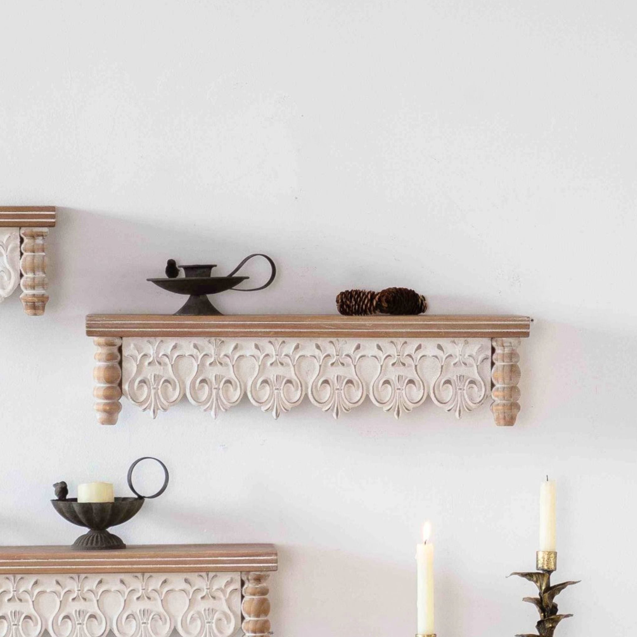 Ariette carved-apron wall shelf, the middle size, styled with a candle holder and pinecones. Hero