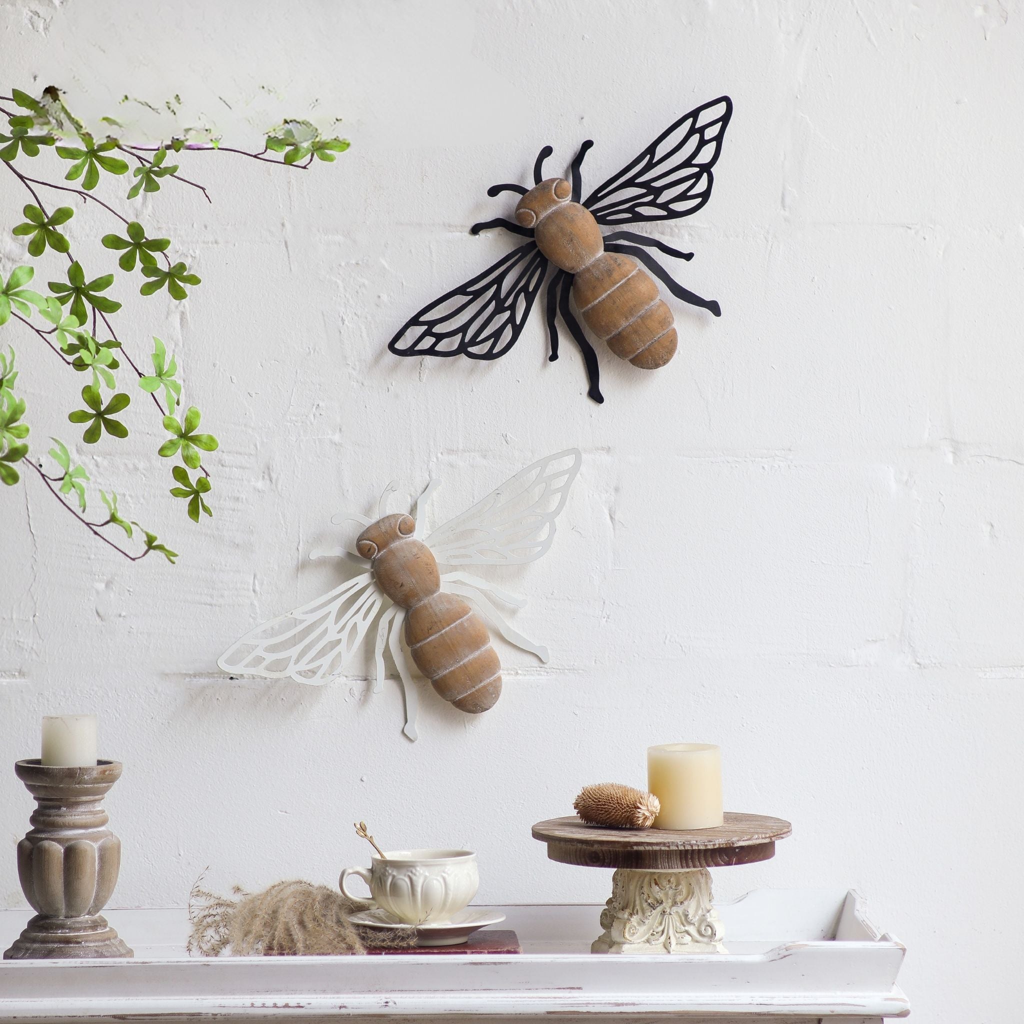 Barry black bee wall decor shown above the antique white Honey bee, demonstrating staggered pair styling against a textured white wall with foliage and styled candles