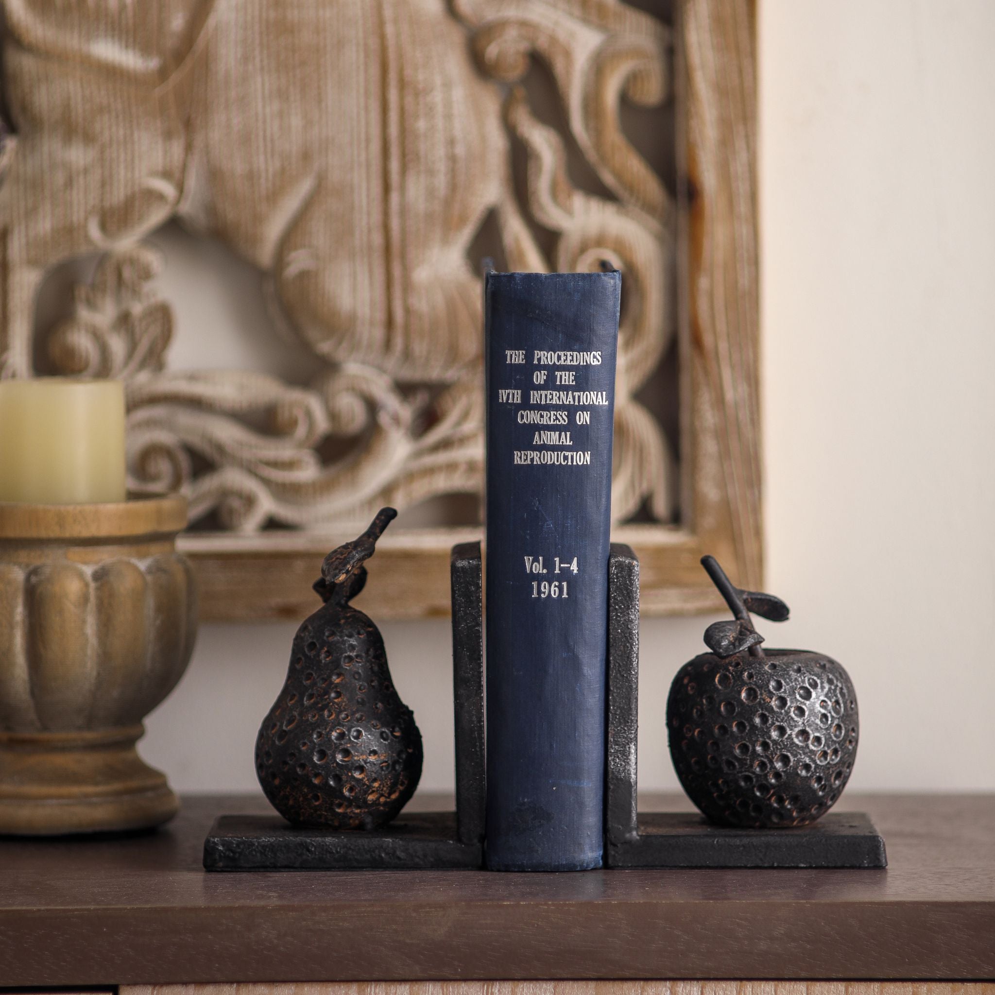 Poire pear and apple bookends holding a navy book on a sideboard with a carved panel behind.