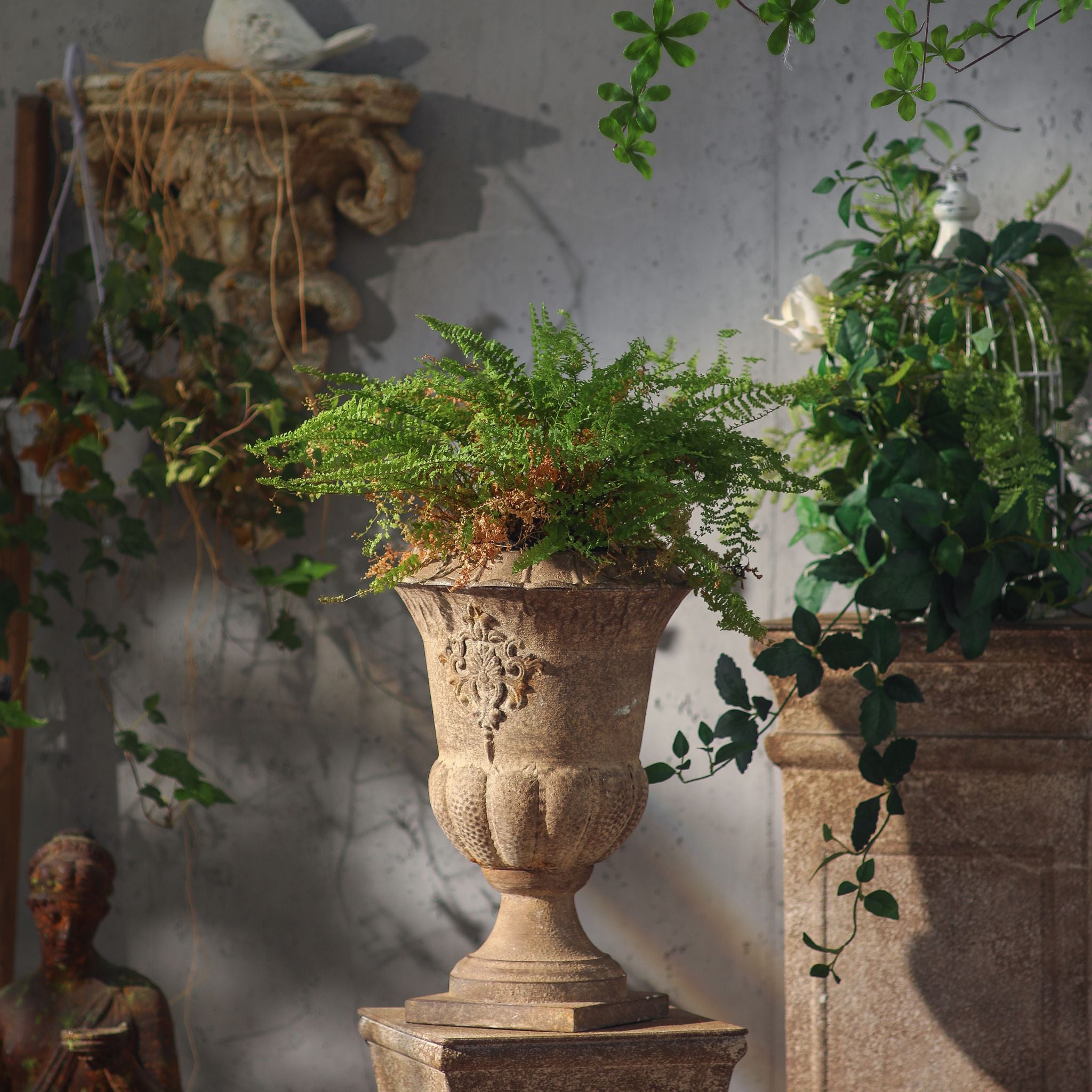Delphine metal urn planted with a fern, styled on a plinth against a grey wall