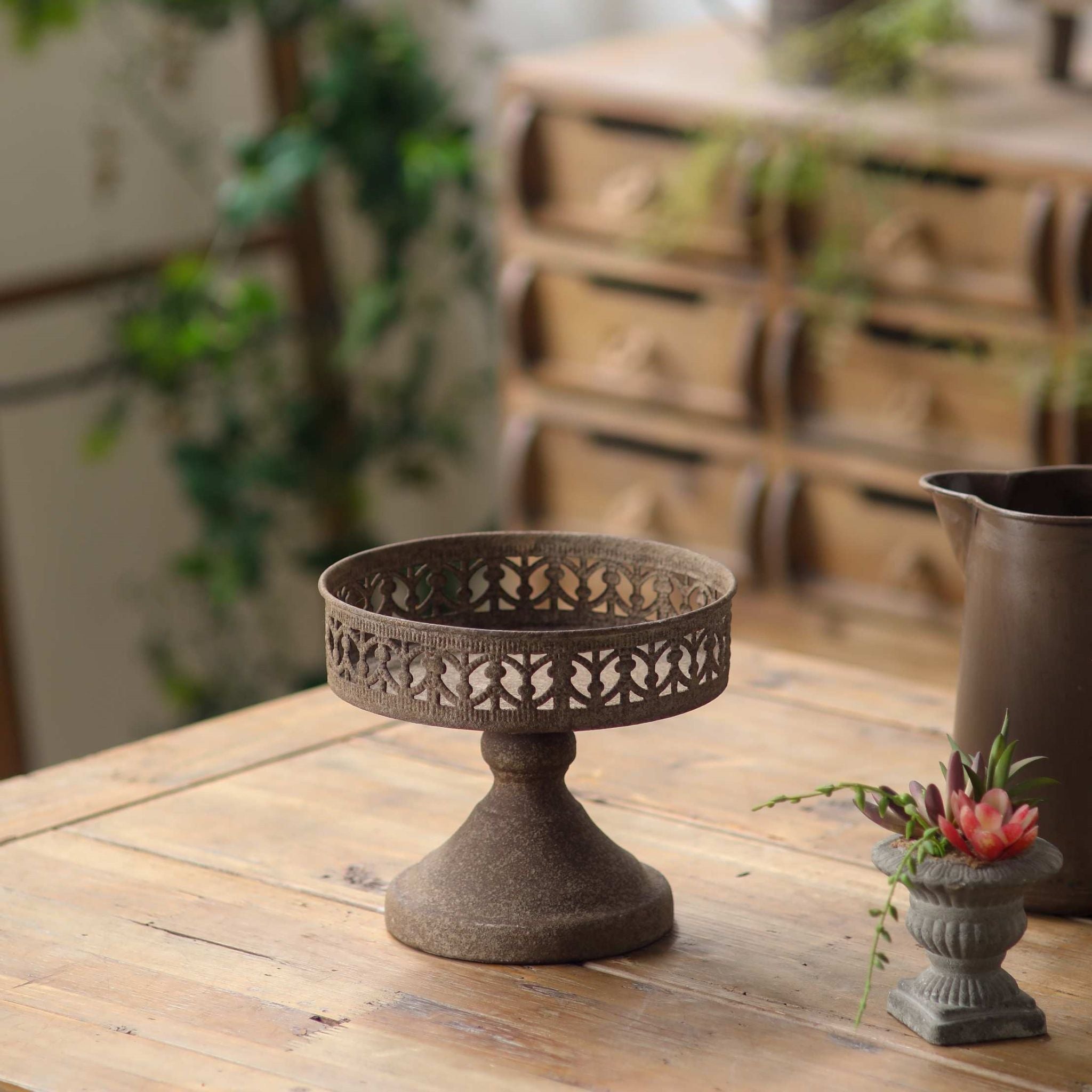 Félice French Provincial pedestal display stand in rustic rust-brown metal with a pierced gallery rim, on a timber table. Hero.