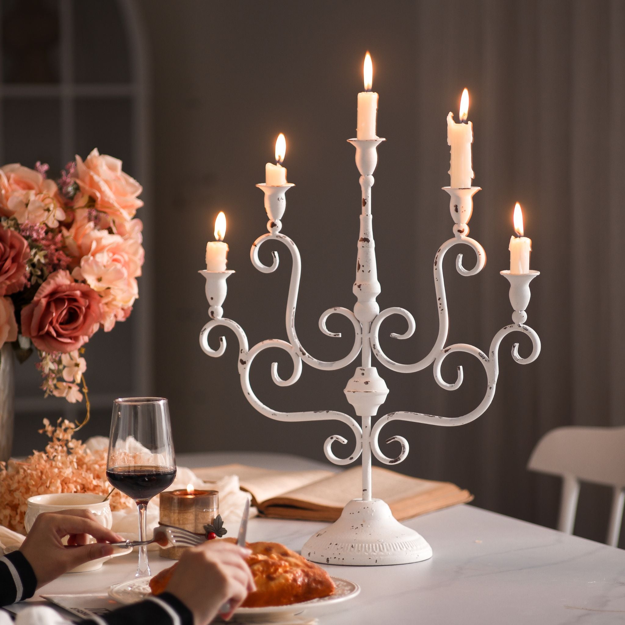 Five-arm candelabra lit on a set dinner table with roses and wine, distressed white finish.