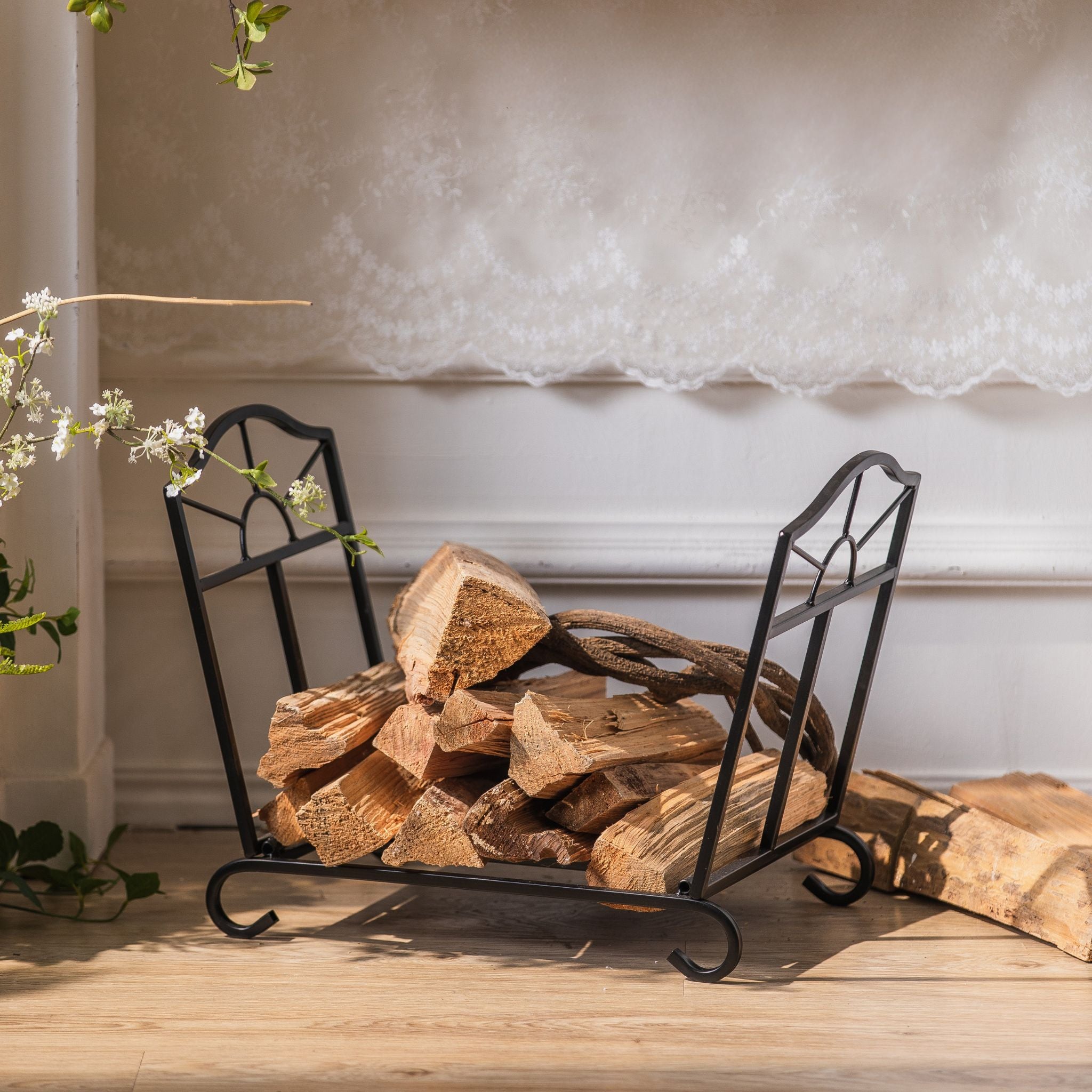 Adrien low matte black firewood log holder with sunburst fan sides, loaded with split logs by a fireplace.