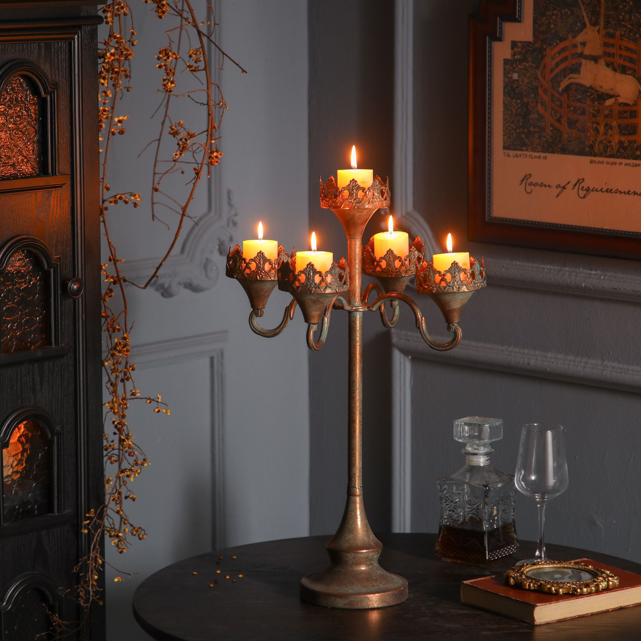 Emma tall five-light antique gold candelabra with crown filigree collars, lit pillar candles, styled on a dark table with a decanter and glass.