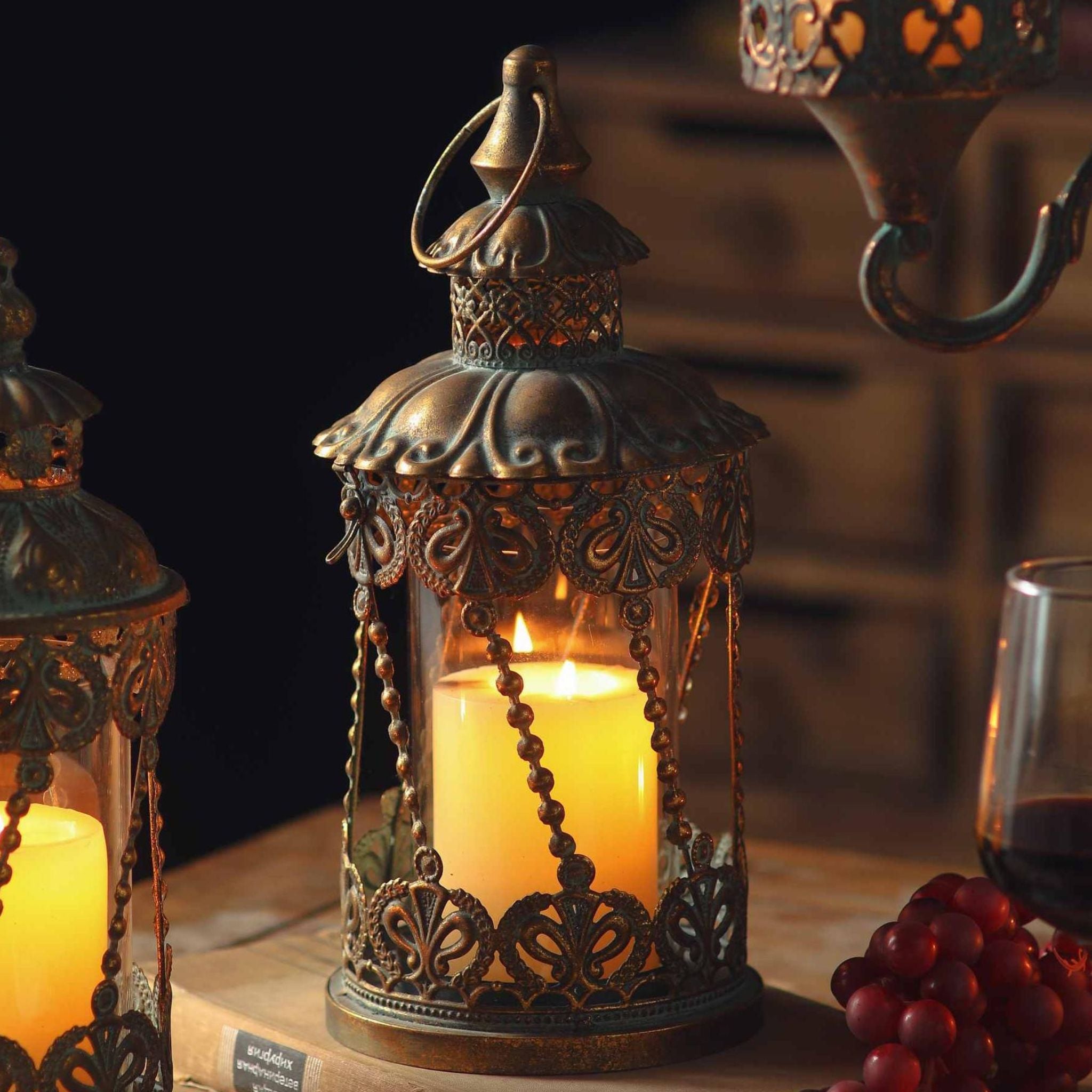 Mael French Provincial filigree lantern in antique gold with a verdigris patina, glass cylinder and lit pillar candle inside, beaded chain detail, on a timber table.
