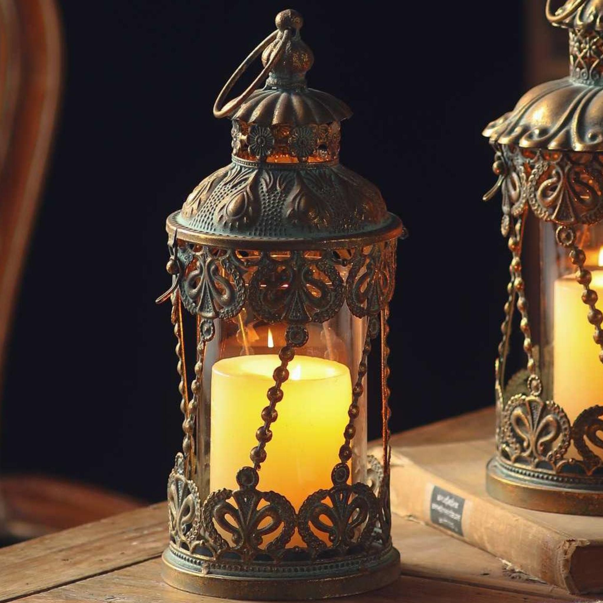 Mylan French Provincial filigree lantern in antique gold with a verdigris patina, glass cylinder and lit pillar candle inside, beaded chain detail, on a timber table.