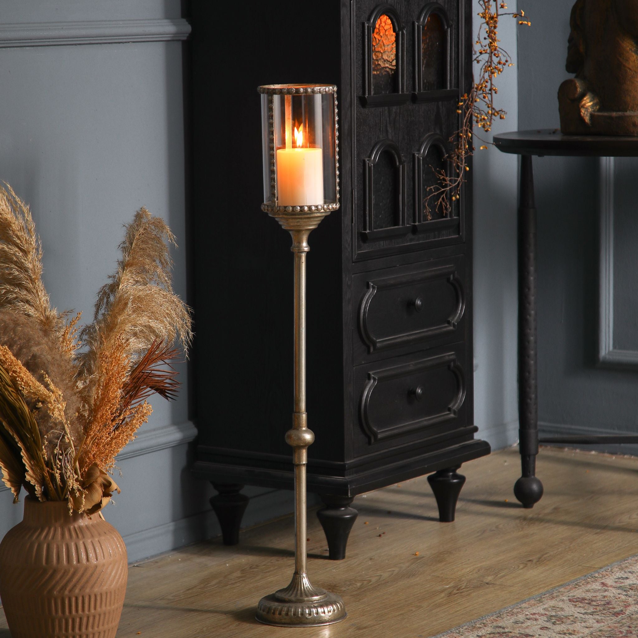 Yves tall antique gold floor candle holder with beaded glass hurricane, lit pillar candle, styled beside a dark cabinet. Hero.