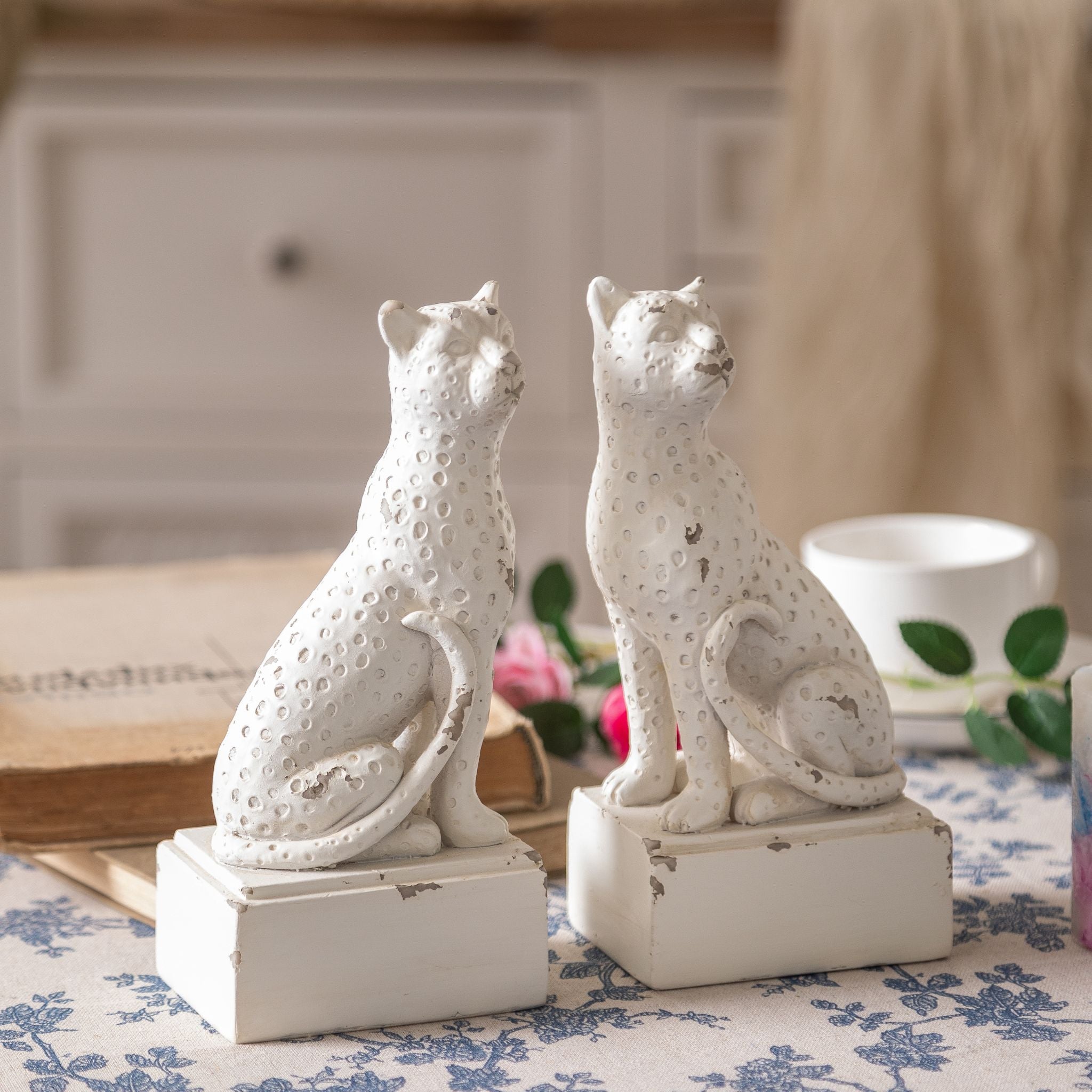 Amandine antique white leopard bookends, a pair of sitting leopard figures on plinths styled on a blue floral cloth