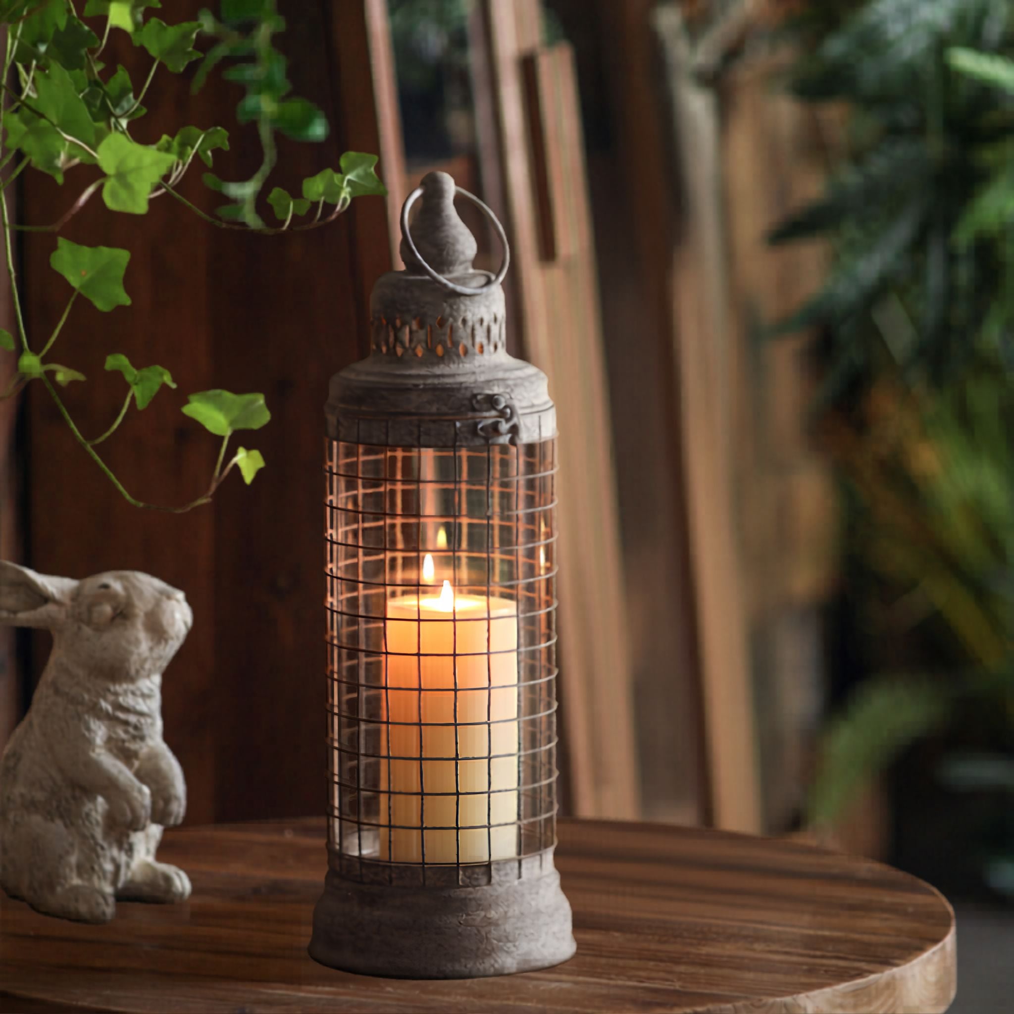 Lyam French Provincial lantern in weathered grey aged metal, wire grid cage over a clear glass hurricane sleeve, lit pillar candle inside, on a timber table.
