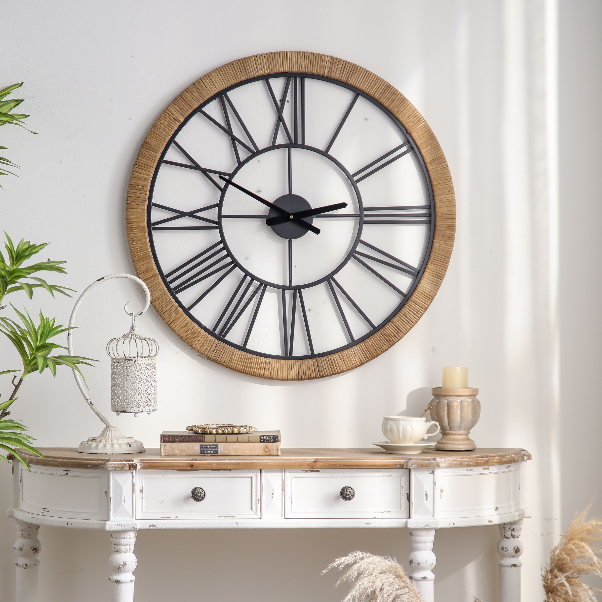 Hugo French Country wall clock with rattan-wrapped frame and black iron Roman numerals mounted above a distressed white console with antique birdcage lantern and styled books. Hero.