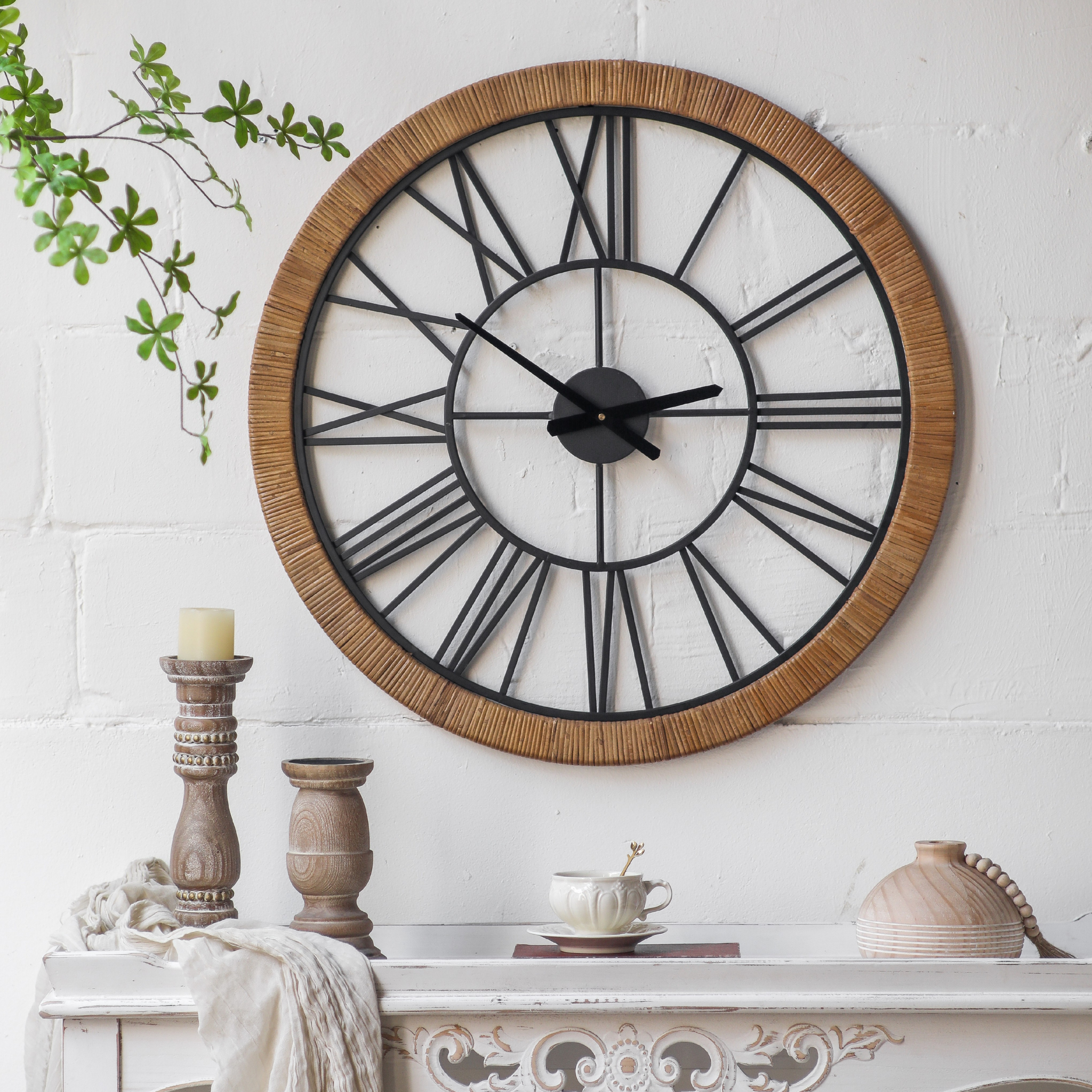 Hugo rattan-wrapped wall clock styled on a mantel above a white console with antique candle holders, teacup, and wood-bead vase