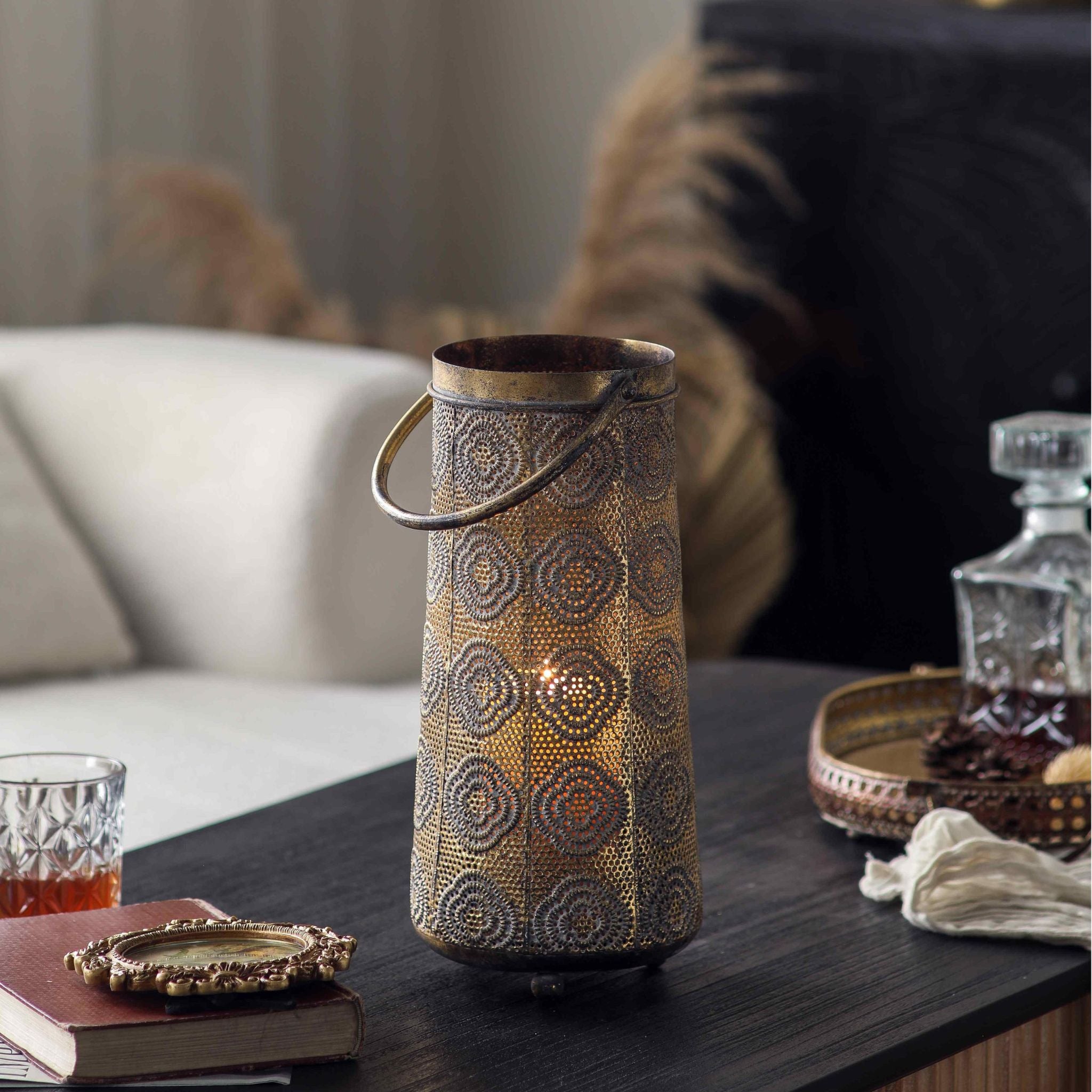 Jasmine pierced metal candle lantern in antique gold, lit on a dark table with the pattern glowing. Hero