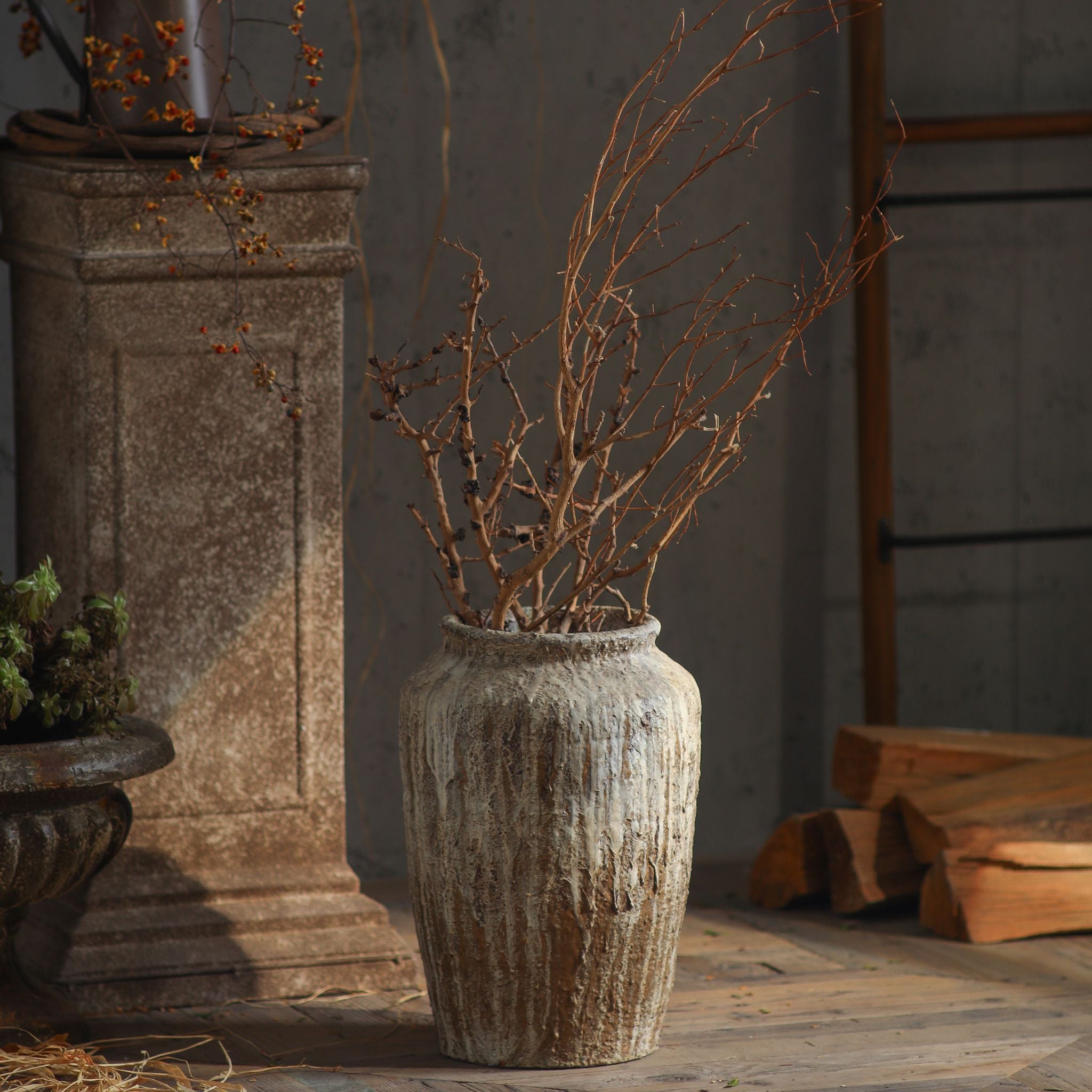 Jean-Marc tall jar vase with a weathered aged stone finish, holding dried branches beside a stone pedestal. Hero.