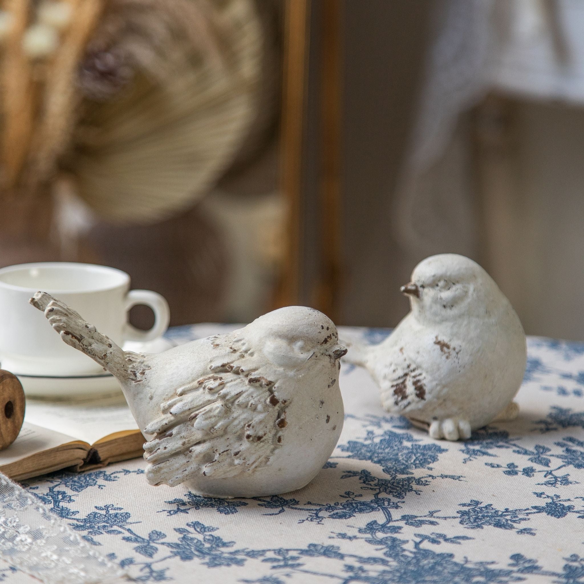 Lucas bird figurines, one with a raised fanned tail on a vintage book and one settled low, showing the worn cream finish