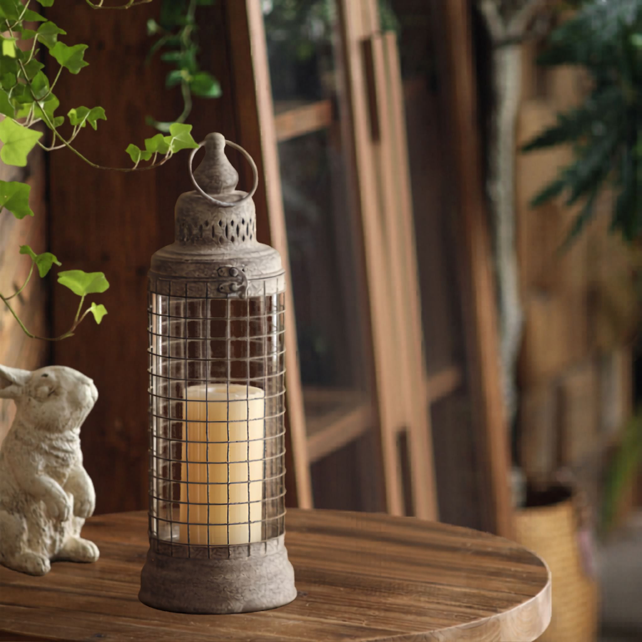 Lyam wire cage lantern in daylight, welded wire grid over a clear glass sleeve with a cream pillar candle inside, on a round timber table.