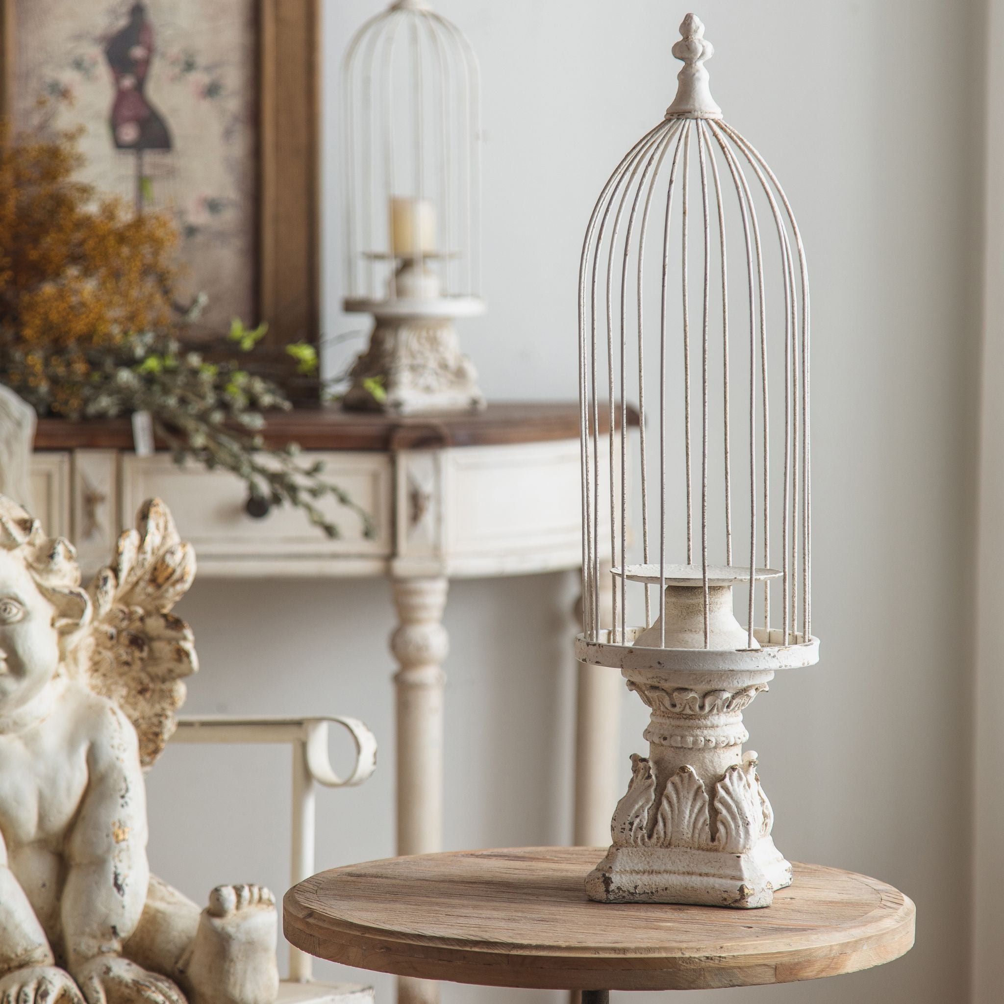 Matheo French Provincial birdcage candle holder in distressed white, domed wire cage on a moulded pedestal, styled on a round table with a cherub and console behind. Hero.