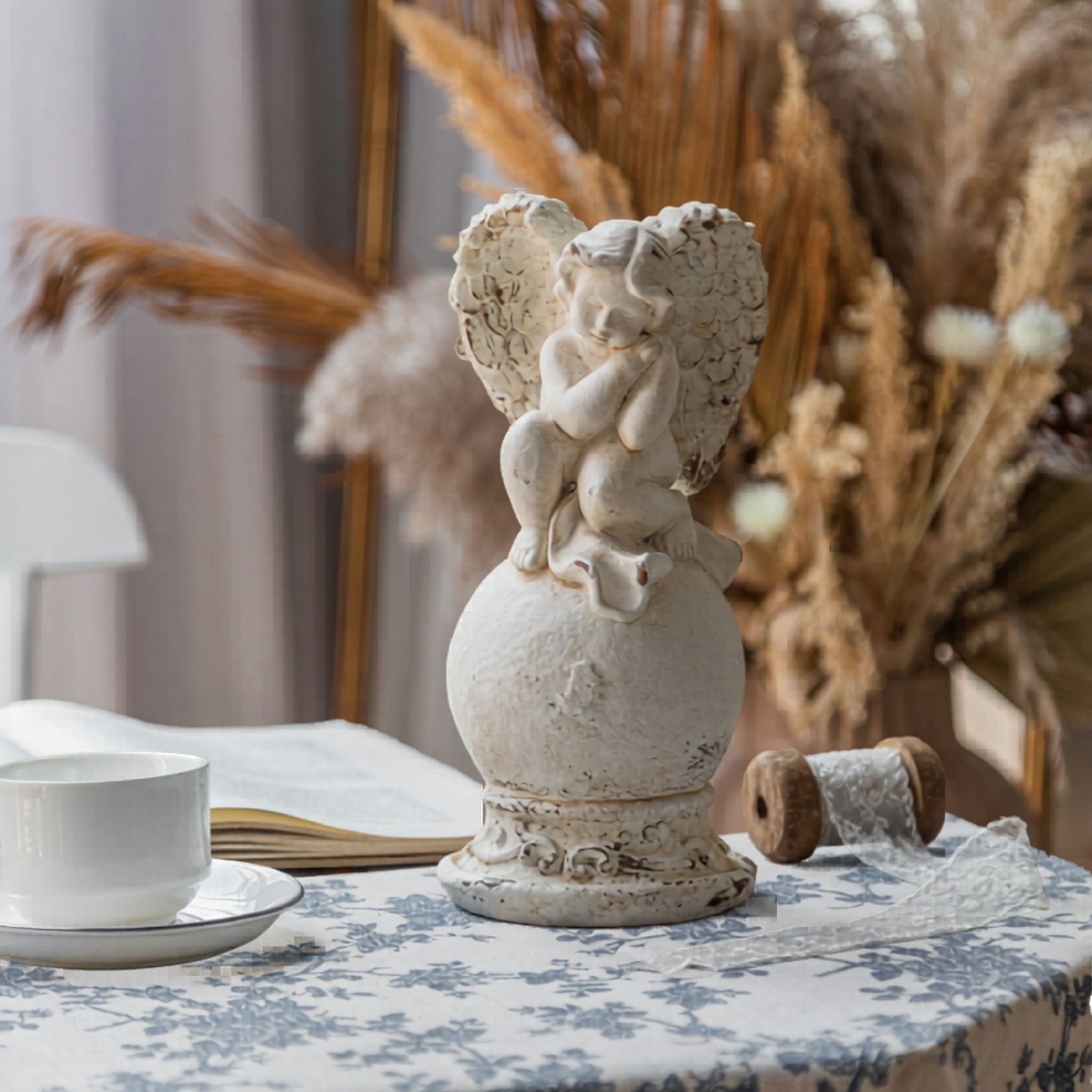 Mathilde French Provincial cherub finial sculpture in distressed whitewash, perched on a scrolled orb pedestal, styled on a blue toile tablecloth. Hero.