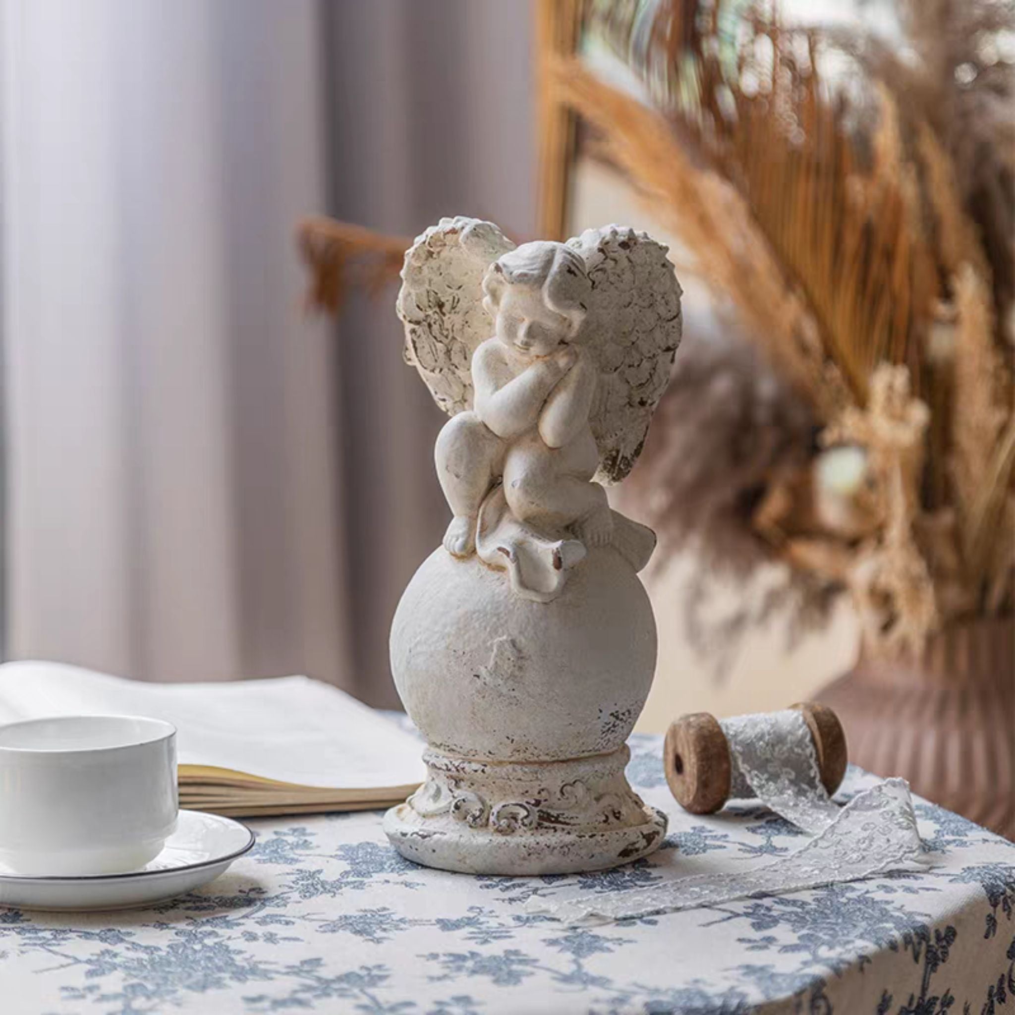 Mathilde cherub finial styled on a blue toile linen with dried pampas grass and lace ribbon