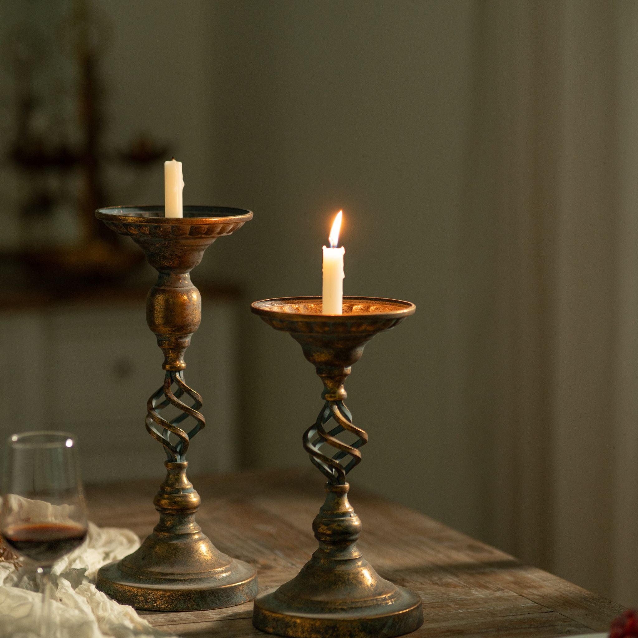 Michel and the shorter Francis candle holders together on a table, one lit, showing the two heights in antique gold and verdigris.