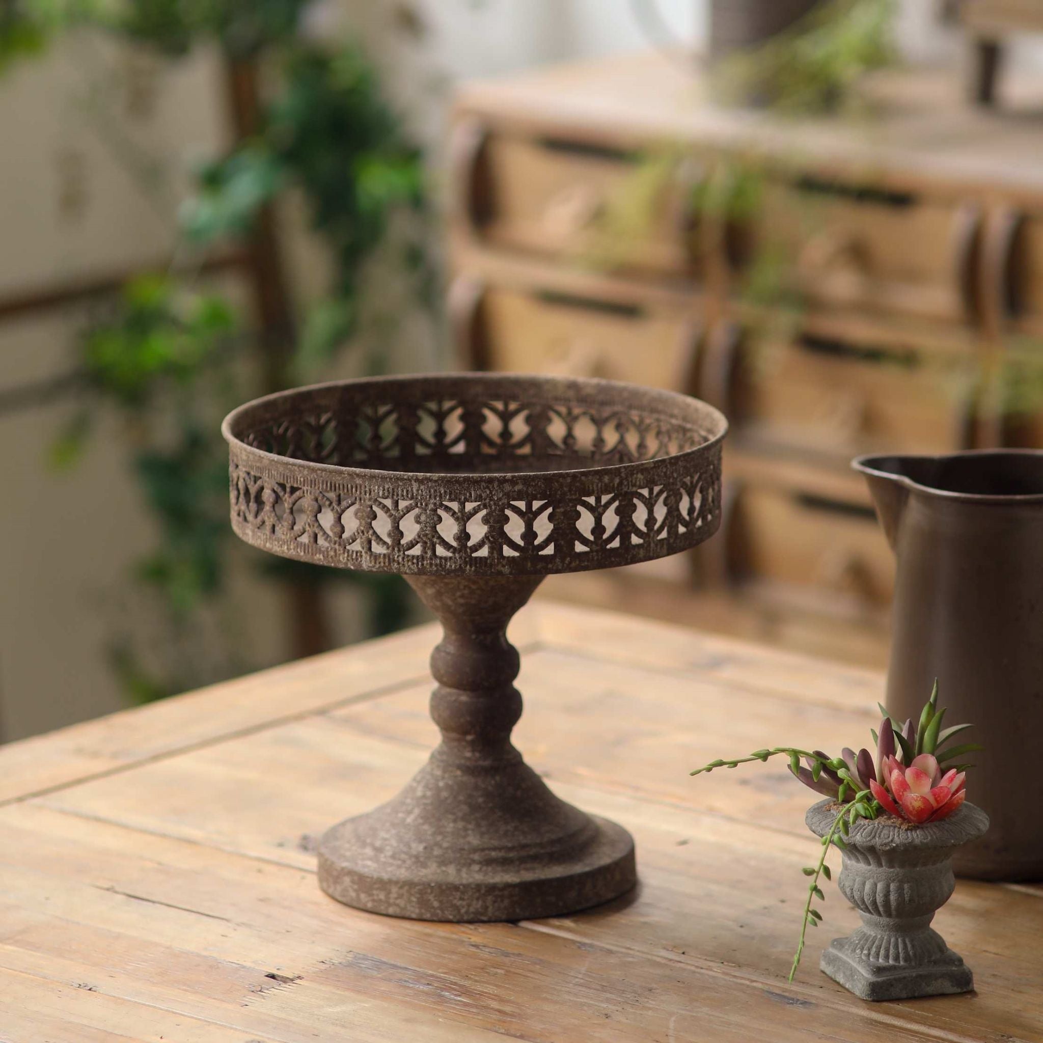 Mirielle French Provincial pedestal display stand in rustic rust-brown metal with a pierced gallery rim, on a timber table. Hero.