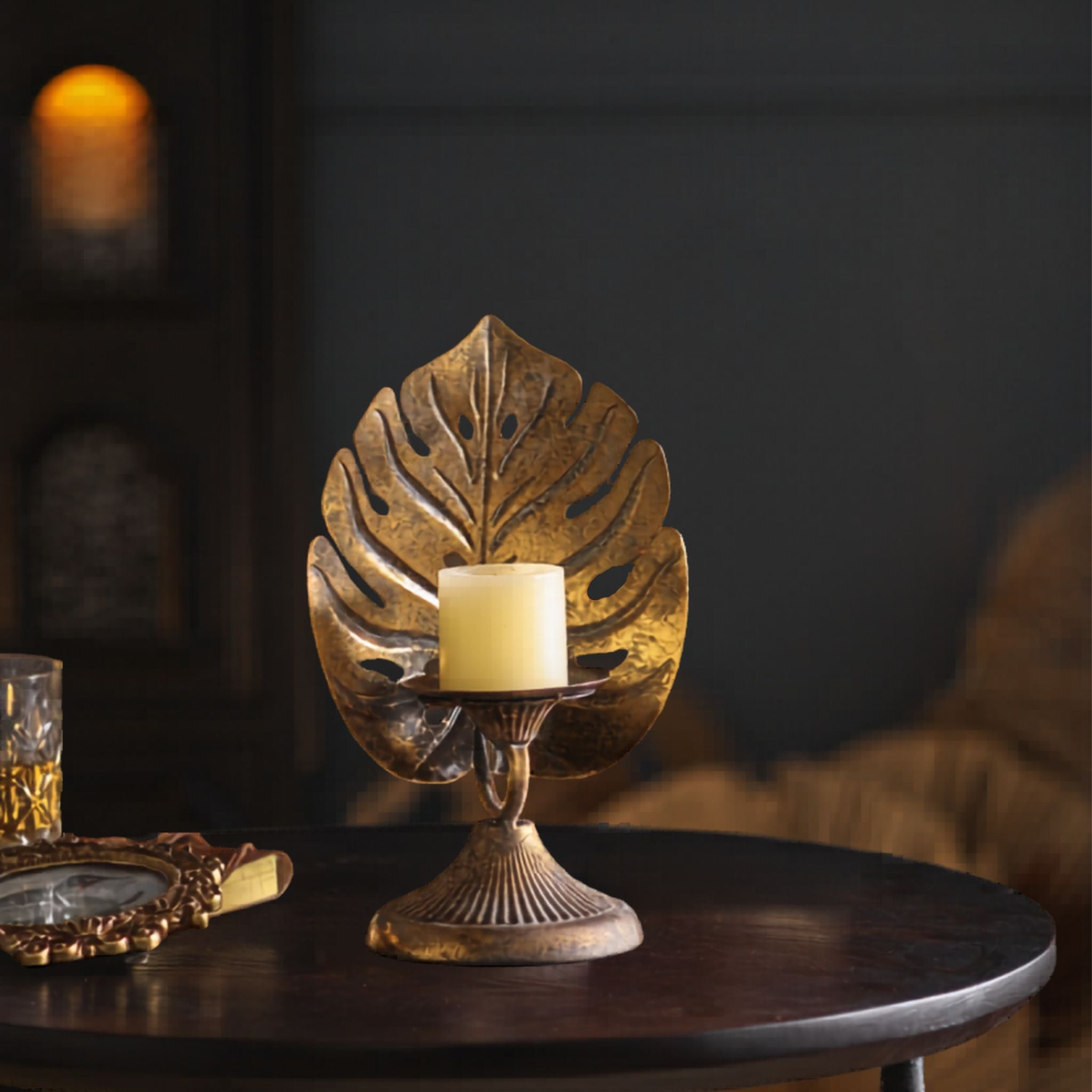 Melanie antique gold monstera leaf pillar candle holder on a round table with a cream candle. Hero.