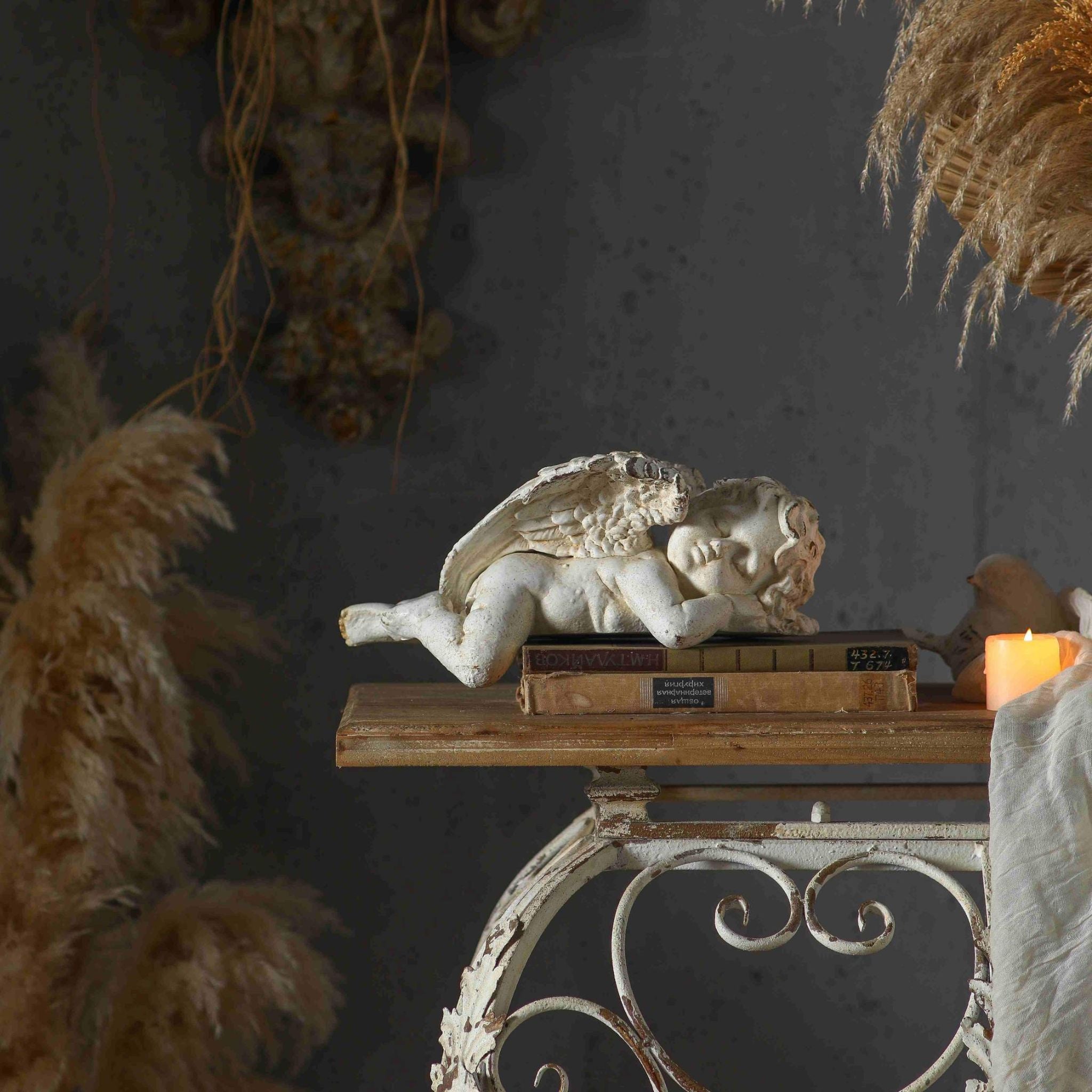 Océane French Provincial sleeping cherub sculpture in heavily distressed white finish, lying on a stack of antique books atop a French country console with iron scrollwork base, styled with pampas grass and candlelight. Hero.