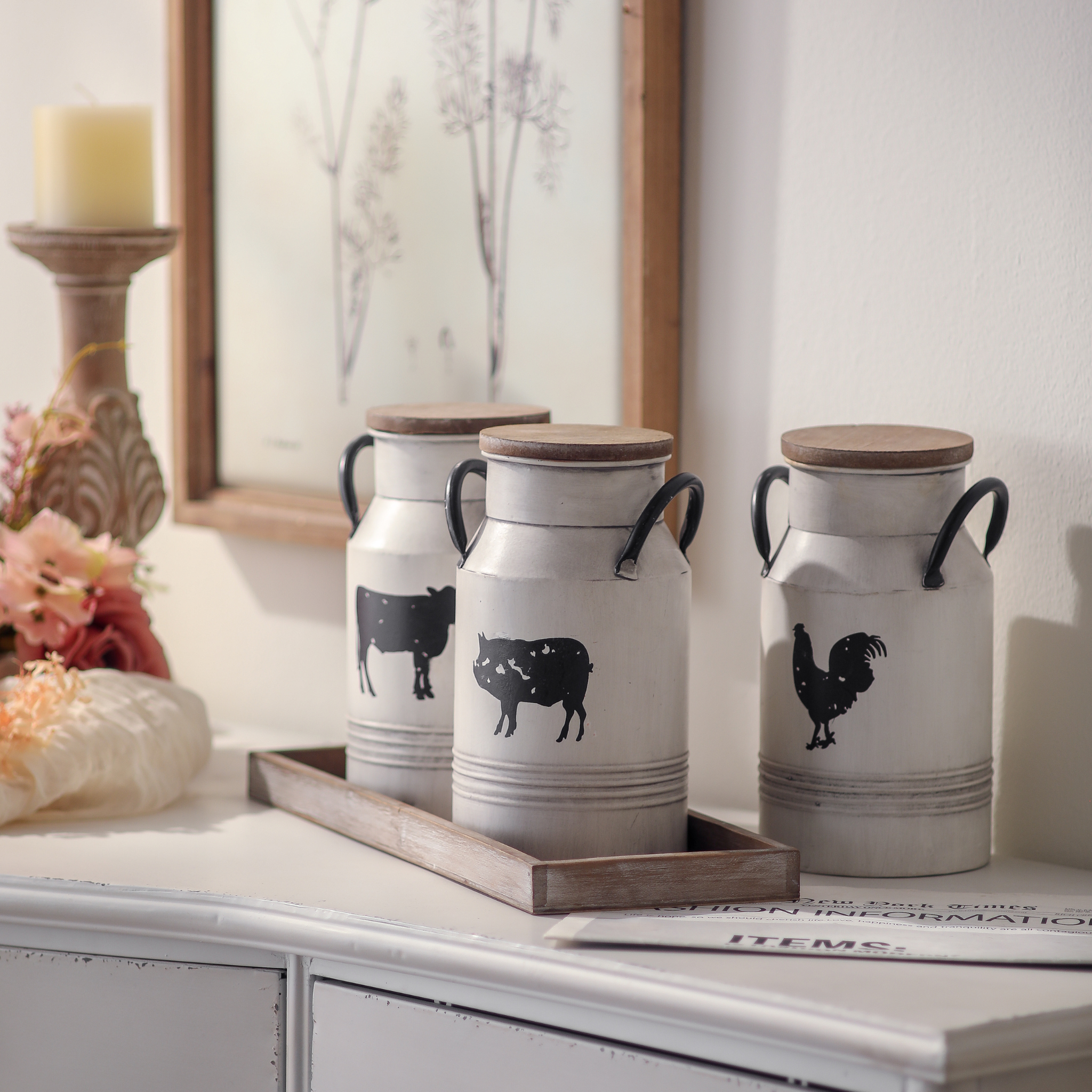 Three farmhouse milk can canisters with cow pig and rooster silhouettes on wooden tray styled with pink florals and pillar candle