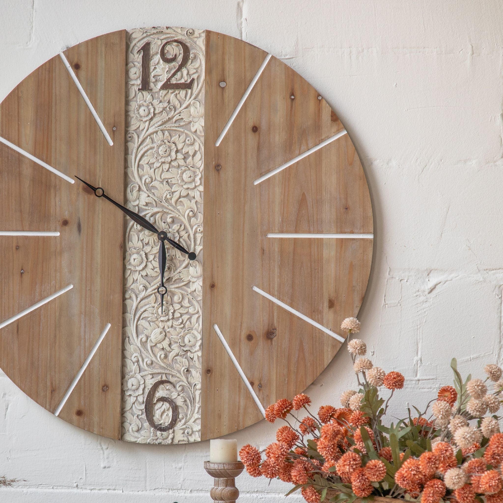 Angled view of Pierre wall clock showing the carved 12 numeral and floral panel in detail, styled with dried orange floral arrangement