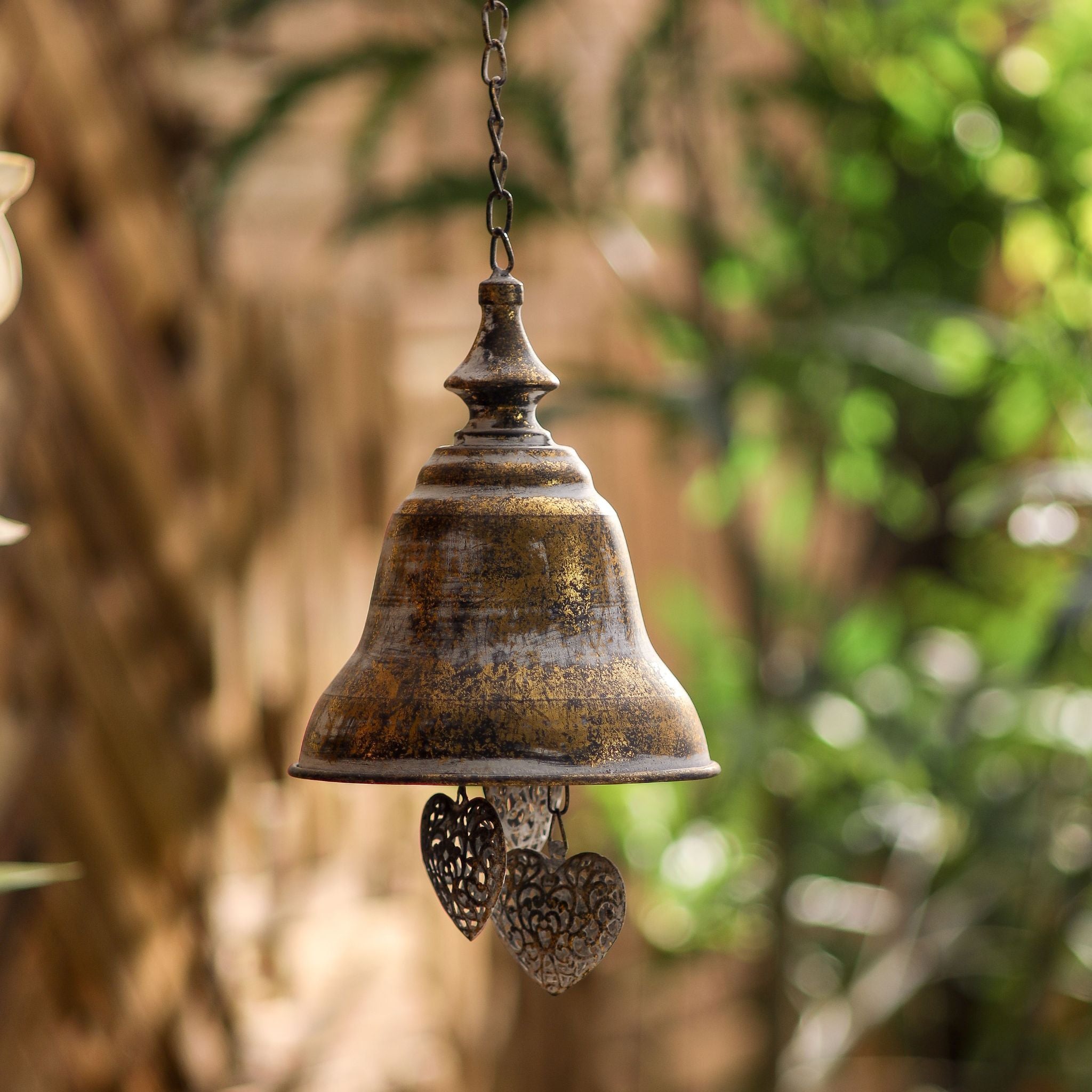 Rochelle French Provincial hanging bell in antique gold metal with pierced filigree hearts, hung in a garden. Hero.