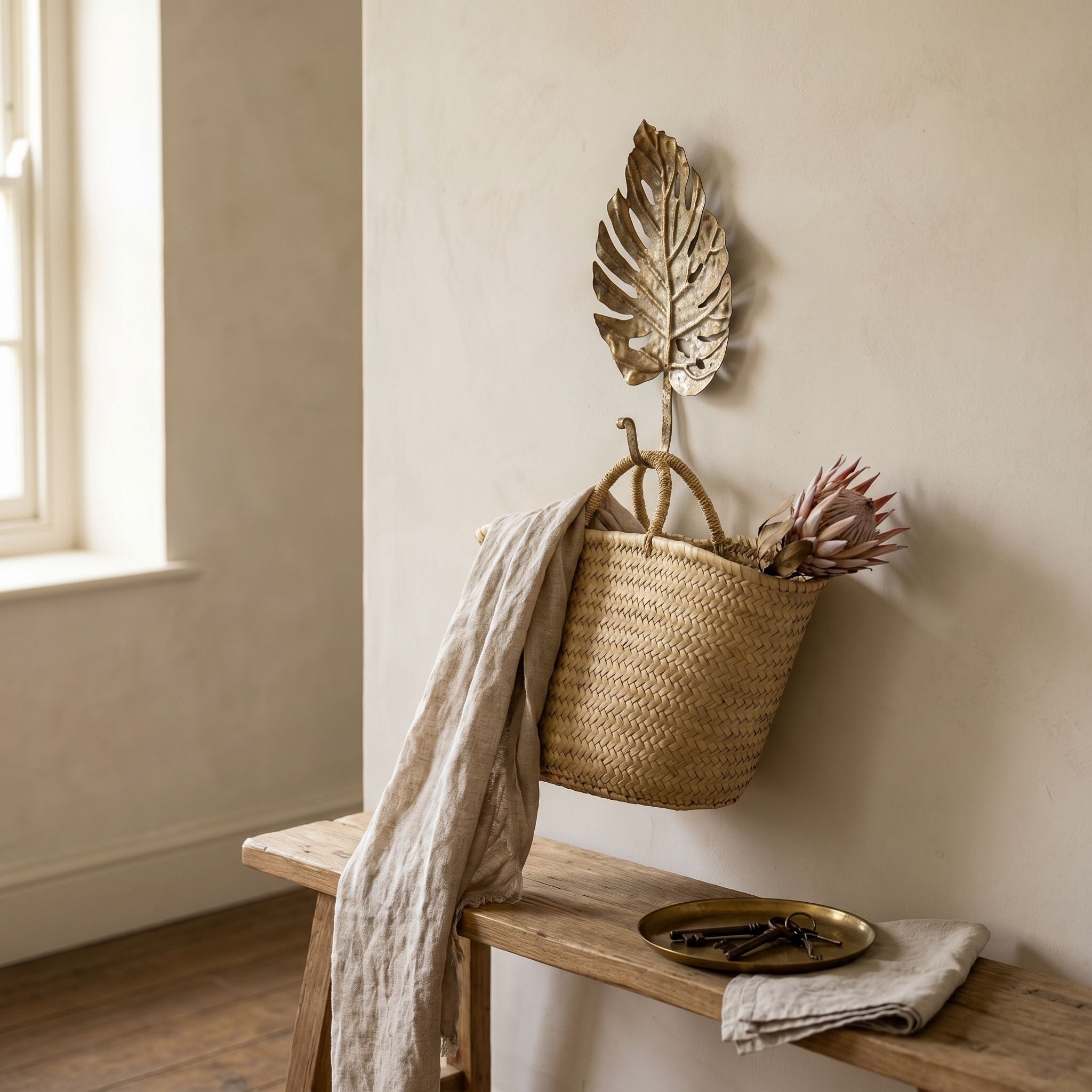 Roxane antique gold monstera leaf wall hook in an entry, holding a woven basket with dried protea and a linen throw above a timber bench.