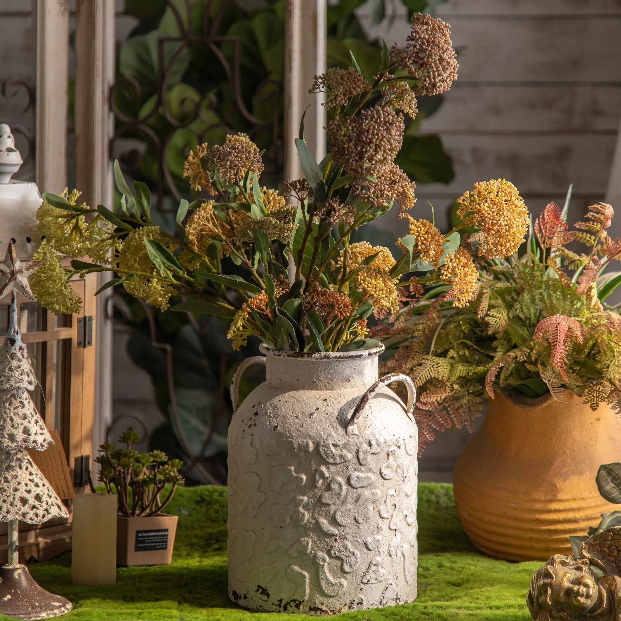 Serge distressed white churn vase with a tall mixed arrangement, beside terracotta pots and greenery.