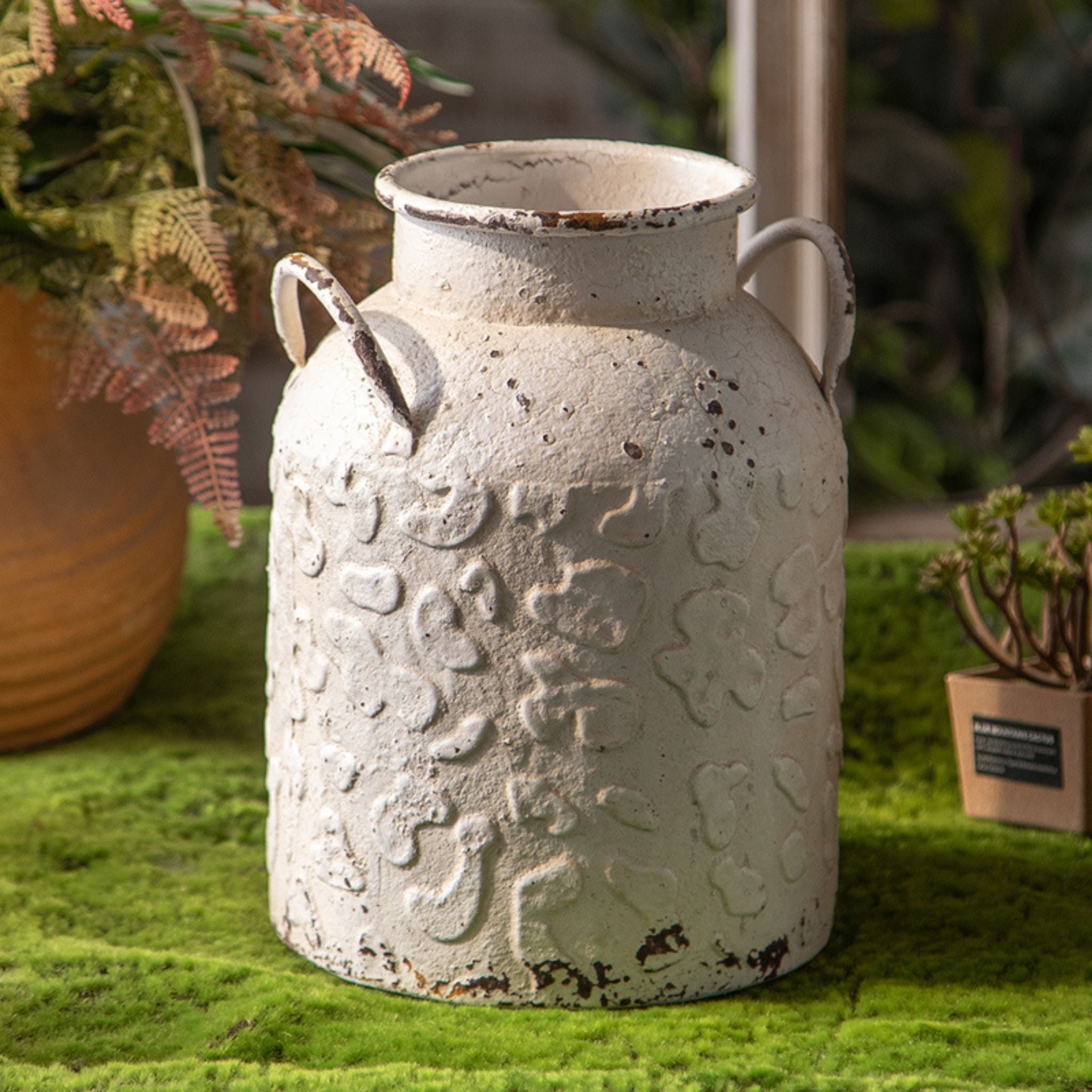 Serge distressed white milk churn vase in embossed metal with twin handles, empty, on green moss.
