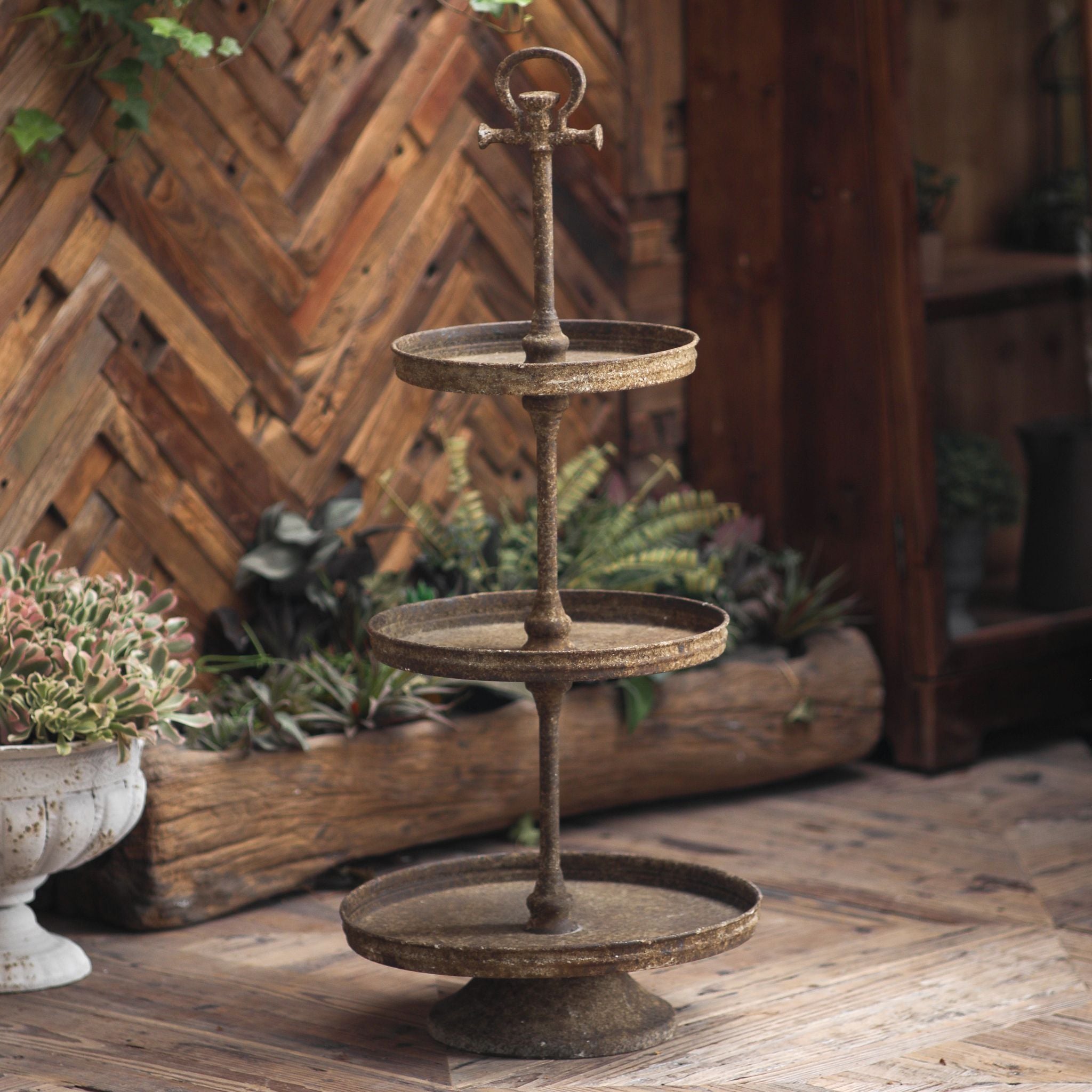 Timéo three tier stand in rustic rust-brown metal, three graduated round trays on a turned pole with a ring finial, on a timber floor. Hero