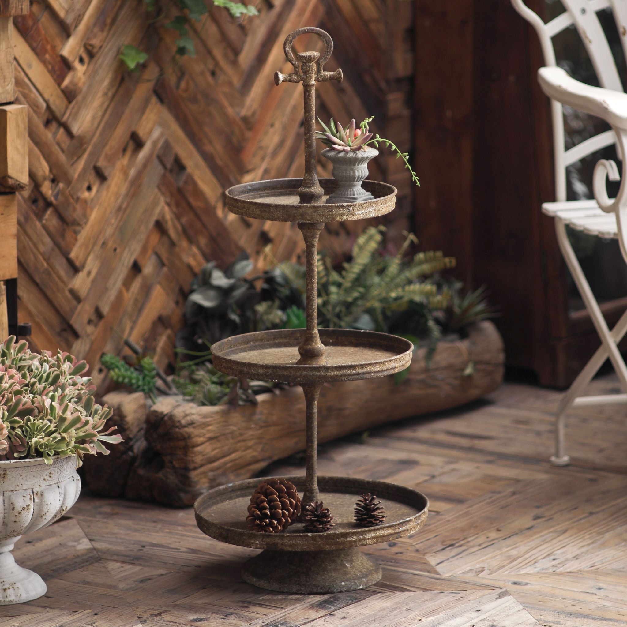 Timéo three tier metal stand styled with a small urn, succulent, and pinecones in a rustic garden setting.