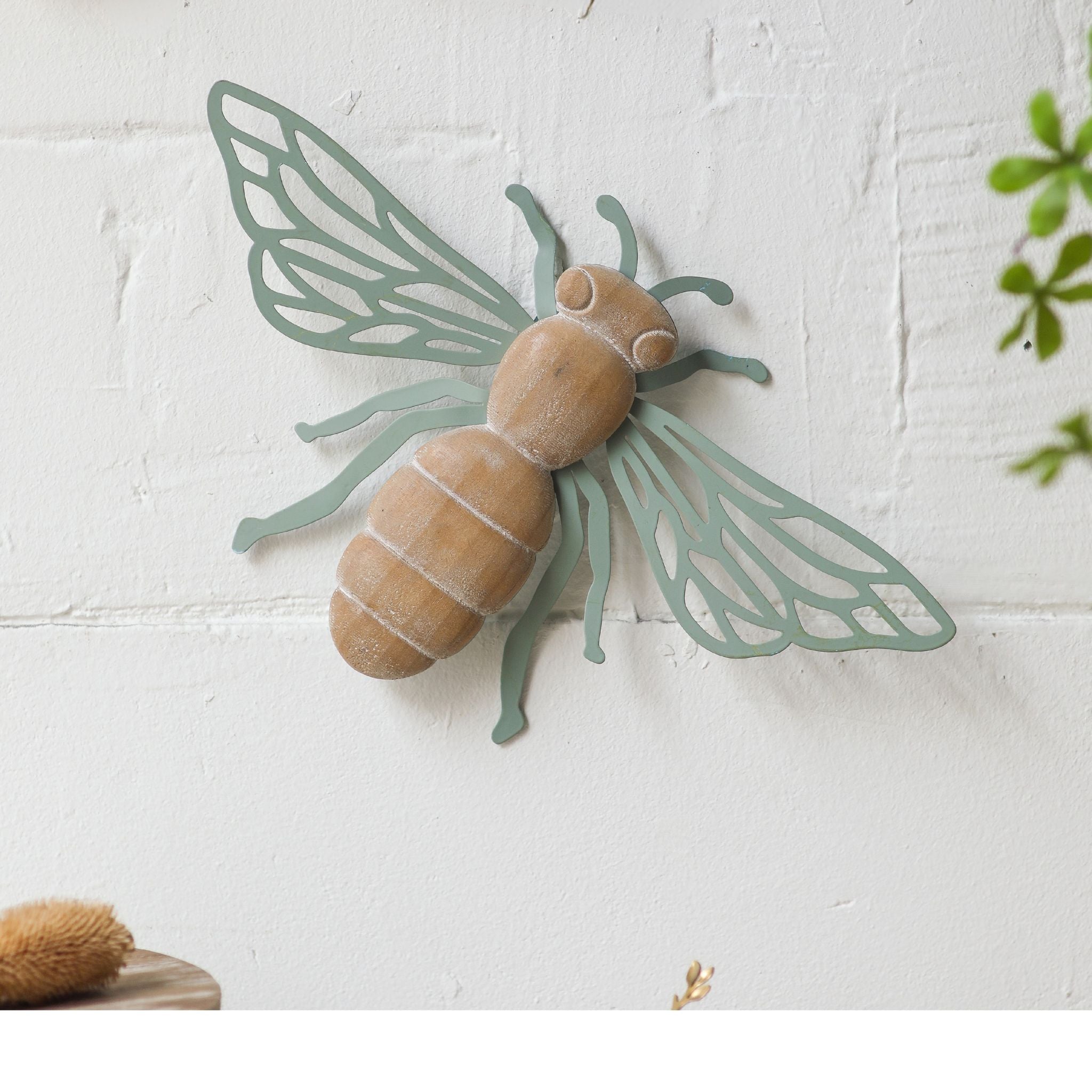 Willy French Provincial bee wall decor with distressed wood body and laser-cut sage green iron wings, mounted solo on a textured white brick wall. Hero