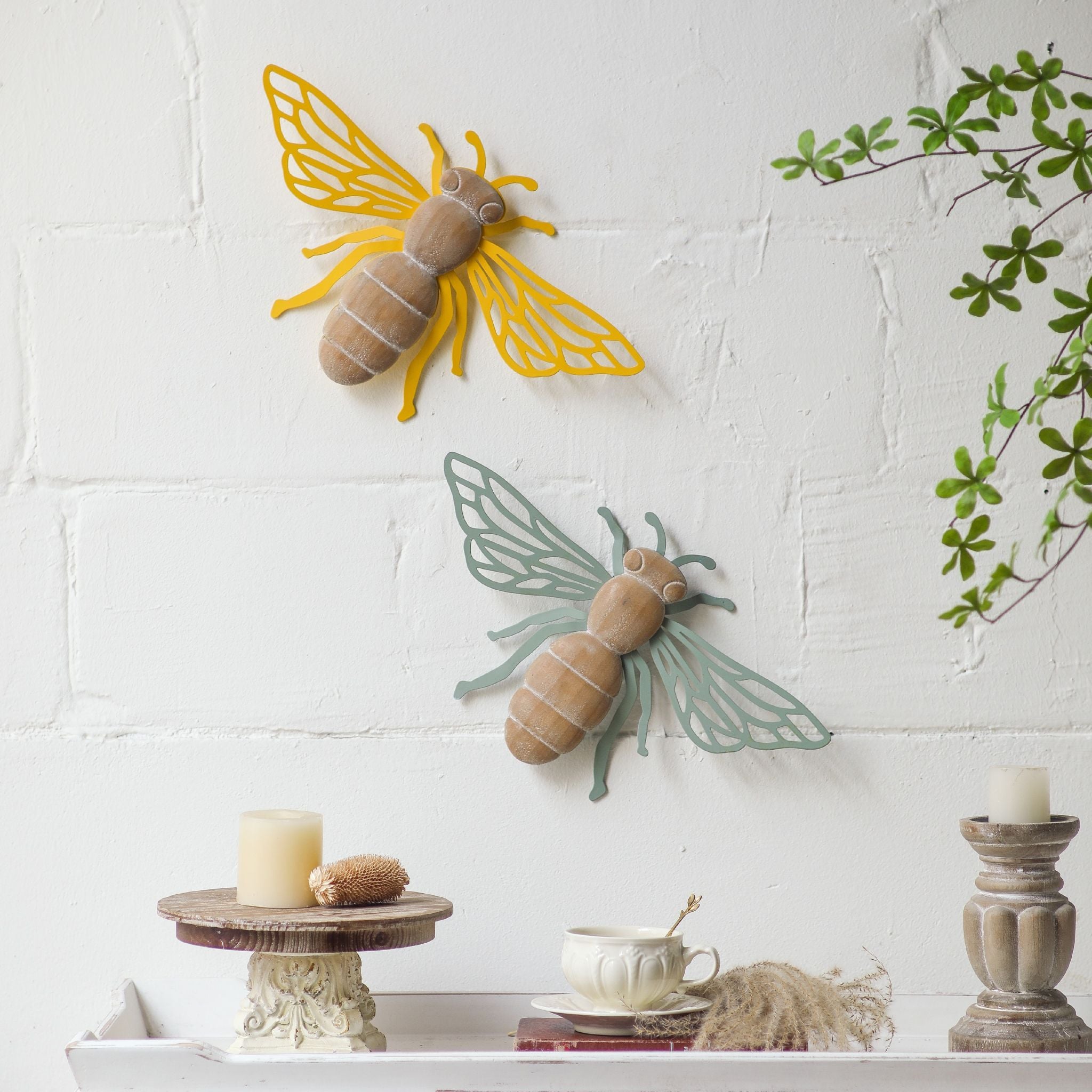 Willy sage green bee wall decor shown below the buttercup yellow Maya bee, demonstrating staggered pair styling against a textured white wall with green foliage