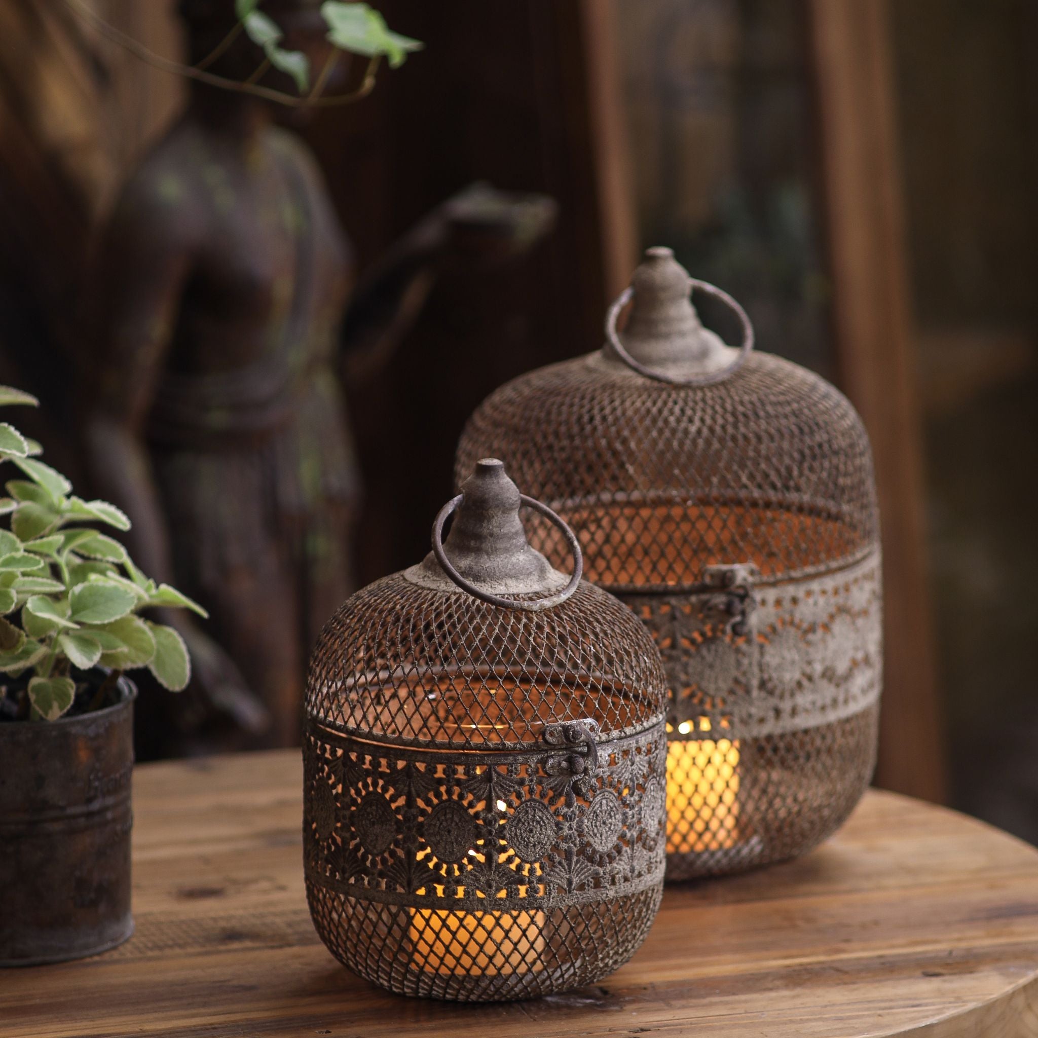 Amani and Afra French provincial lanterns, distressed aged iron with pierced floral band and woven mesh dome, glowing candlelight on a rustic timber table.