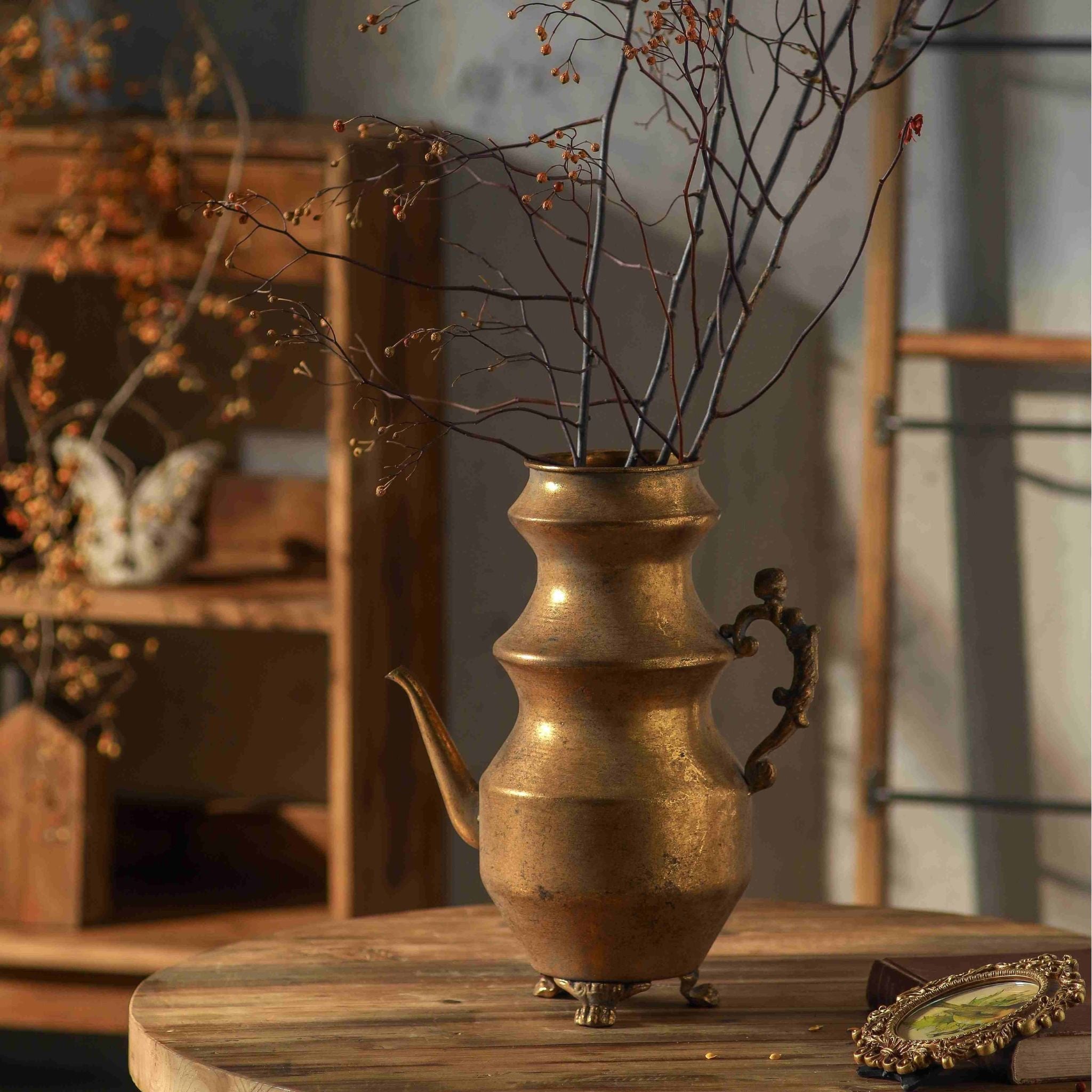 Ivo teapot vase in antique gold holding dried branches on a round timber table